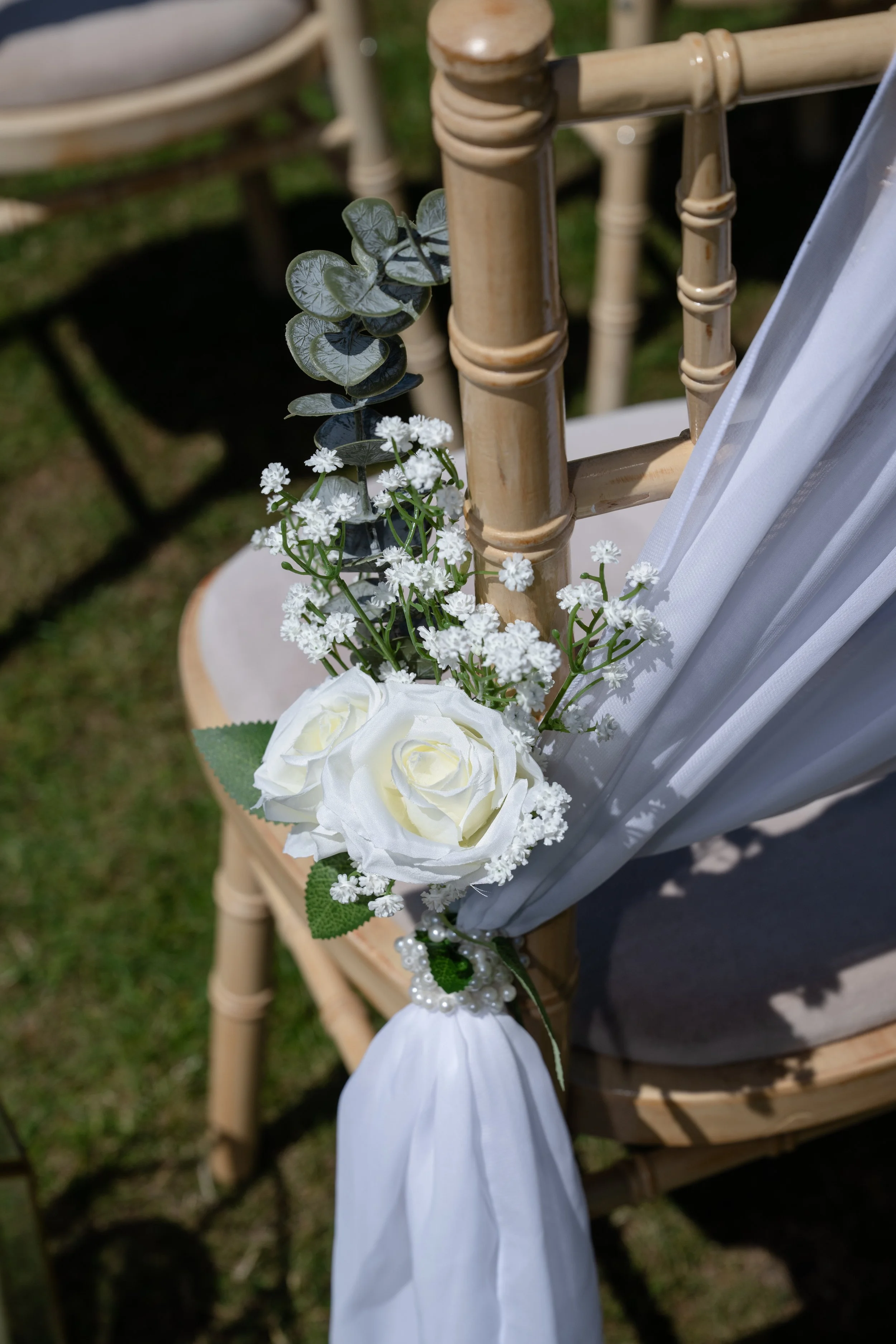 Close-up of a decorated beige wooden chair with white fabric cash sash, artificial white roses, small white flowers, and green leaves attached to the backrest, ideal for an outdoor ceremony.
