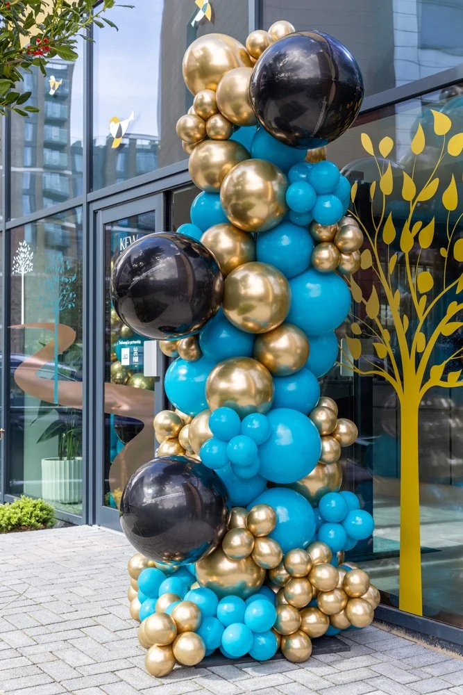 Decorative balloon arrangement with gold, black, and blue balloons outside a modern glass building with a painted yellow tree design on the window. Themed event planning and accessories. 