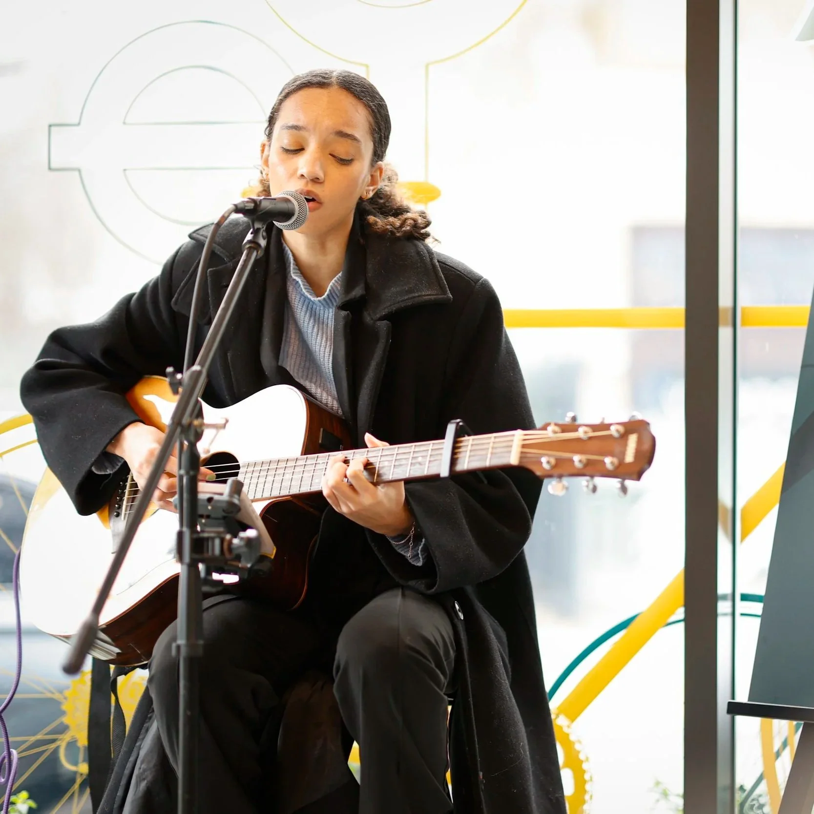 Young woman with curly hair playing an acoustic guitar and singing into a microphone indoors. Event entertainment and planning.