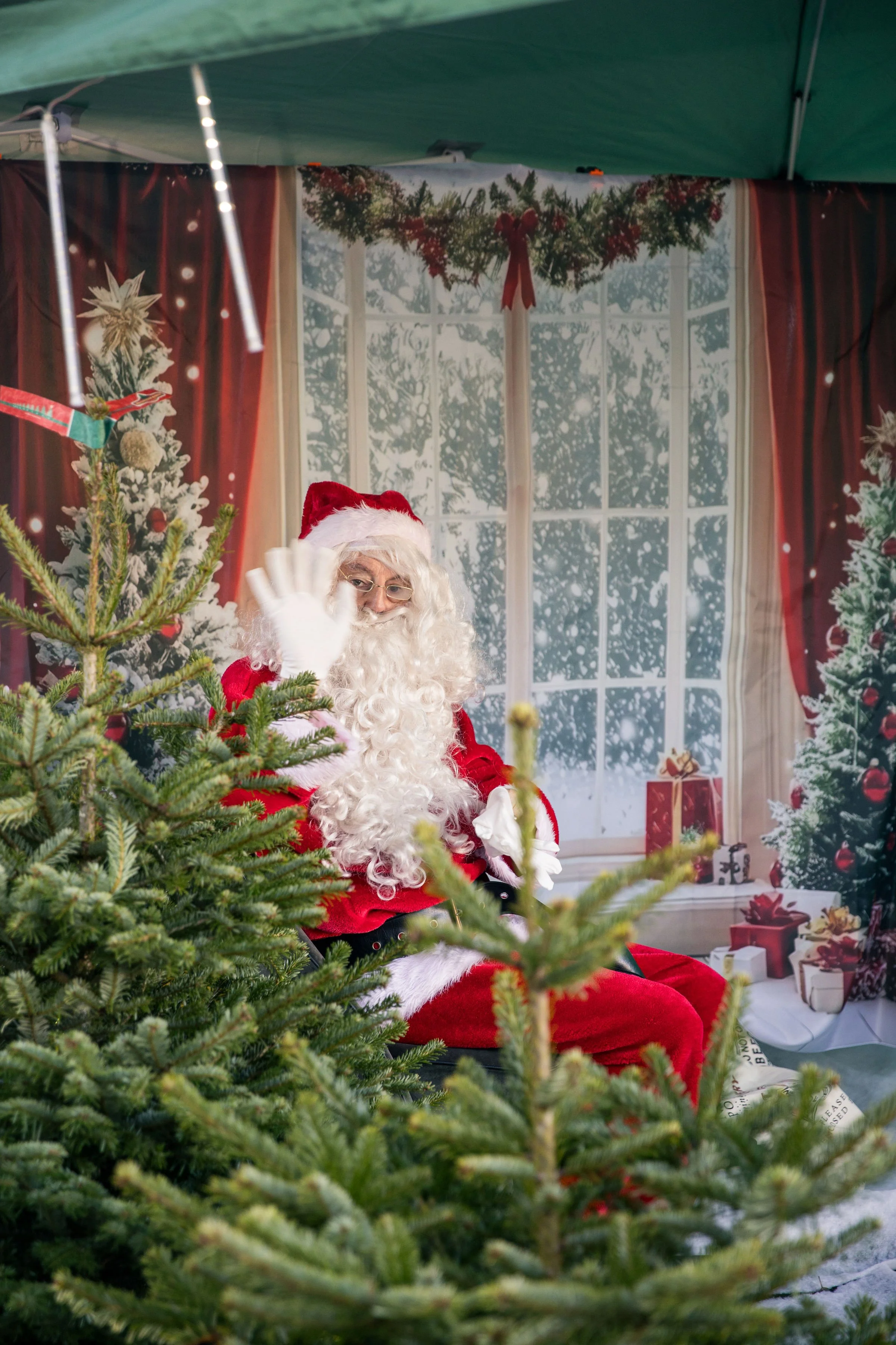 Santa Claus sitting in front of a Christmas backdrop, waving, with decorated Christmas trees in the foreground. Themed event planning.