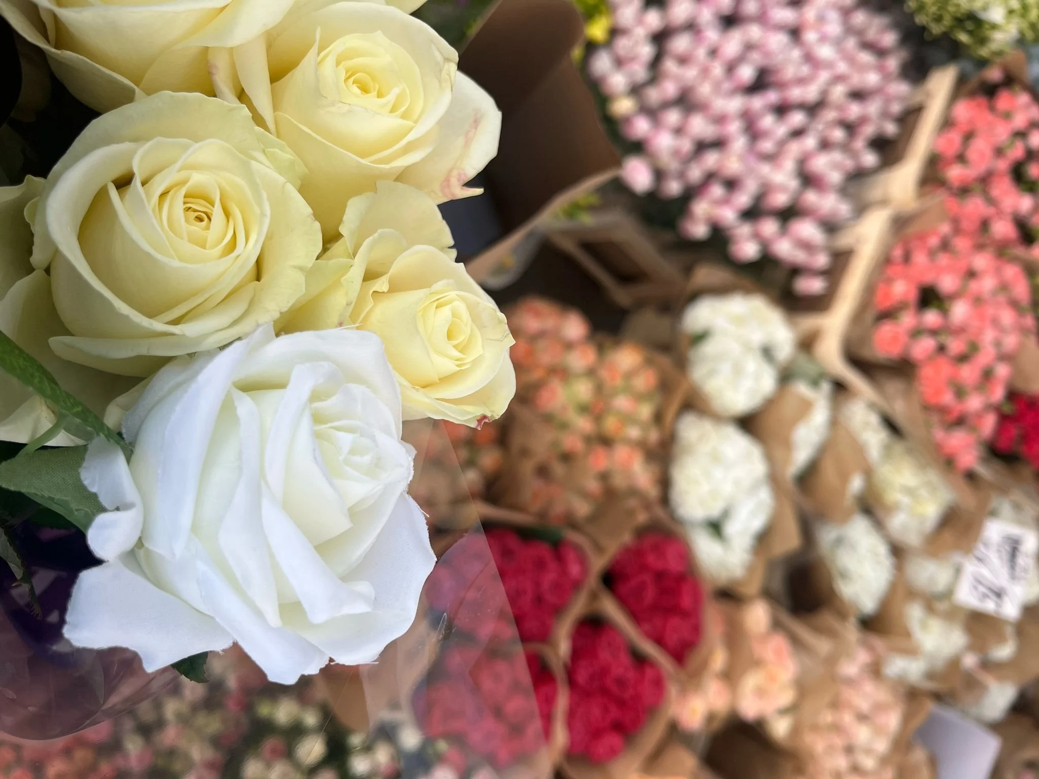 A bouquet of white and pale yellow roses in the foreground with various other flower arrangements in pots and containers in the background.