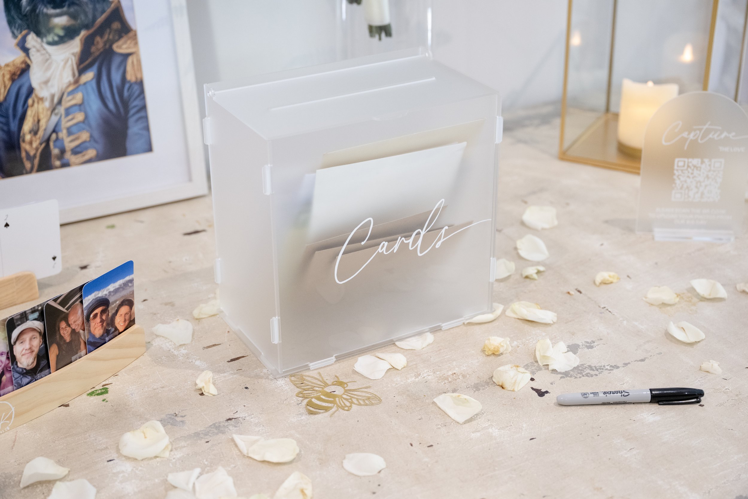 A transparent card box labeled 'Cards' on a table with scattered white rose petals, a Sharpie marker, framed photos, and decorative items. Personalised event stationery for weddings and events.