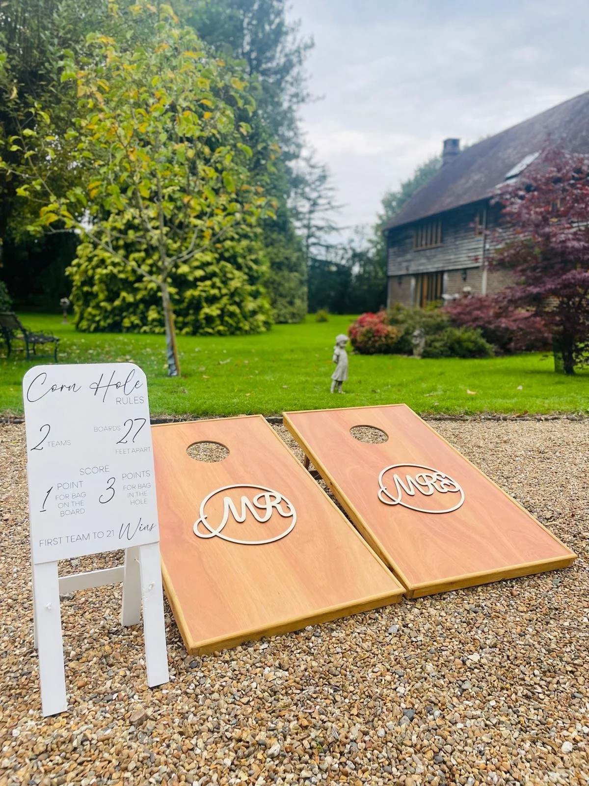 Cornhole game setup on a gravel surface outside, with a whiteboard listing rules, and a lush backyard with trees and a house in the background. Forms part of a Giant Lawn Games package.