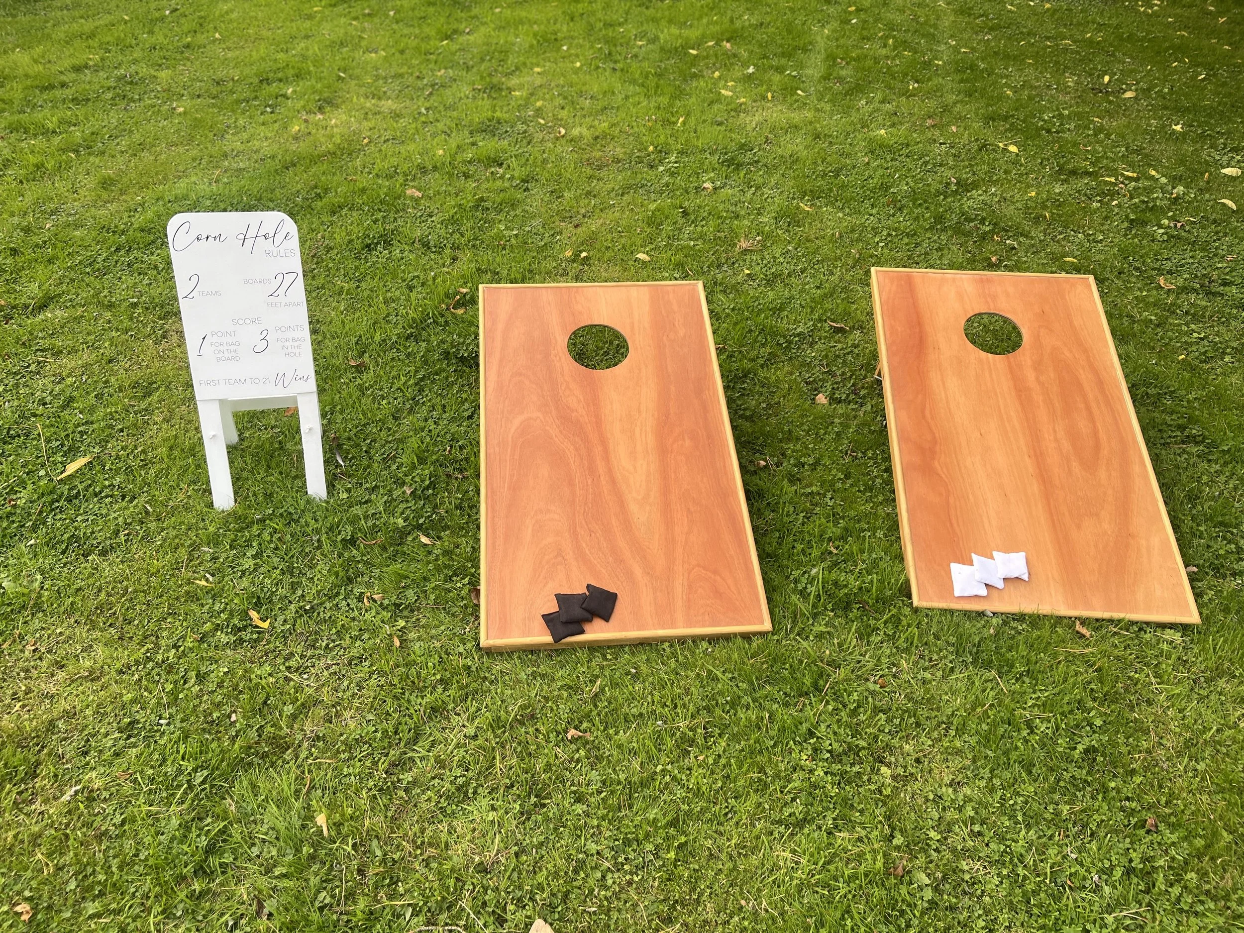 Two cornhole game boards with black and white bean bags on a grassy lawn, and a white sign with rules for the game.
