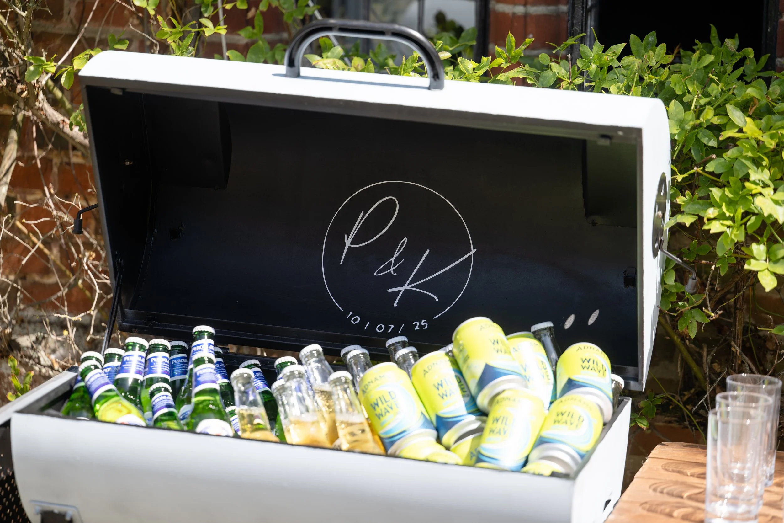 An outdoor BBQ filled which has been personalised with a wedding logo. Filled with green and blue bottles and yellow cans of beverage, set against greenery and a wooden table with empty glasses.