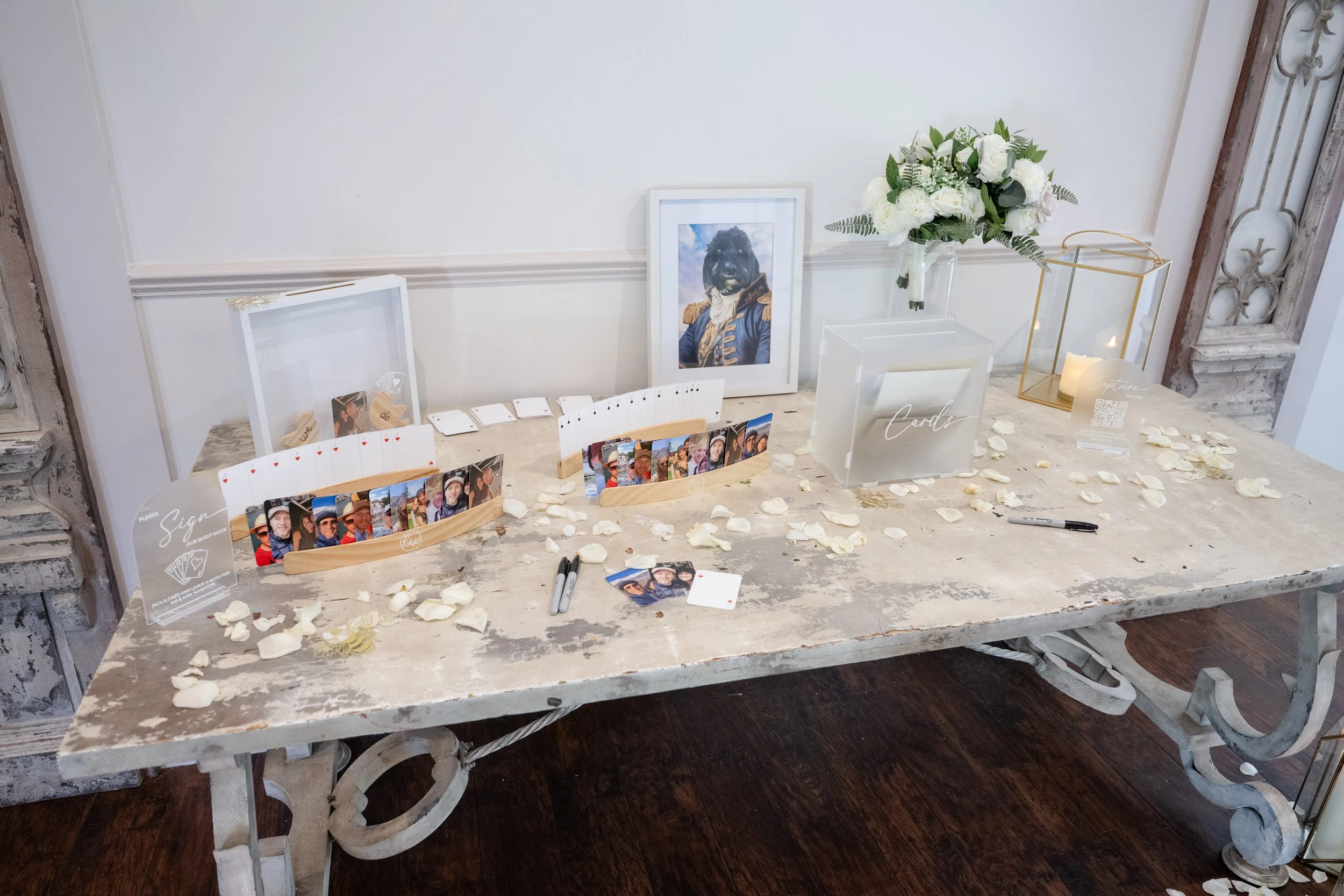 Table decorated with photo displays, a framed portrait of a dog in historical costume, white rose petals, candles, and signages for guest signatures and cards. Personalised event stationery and accessories for weddings &events.