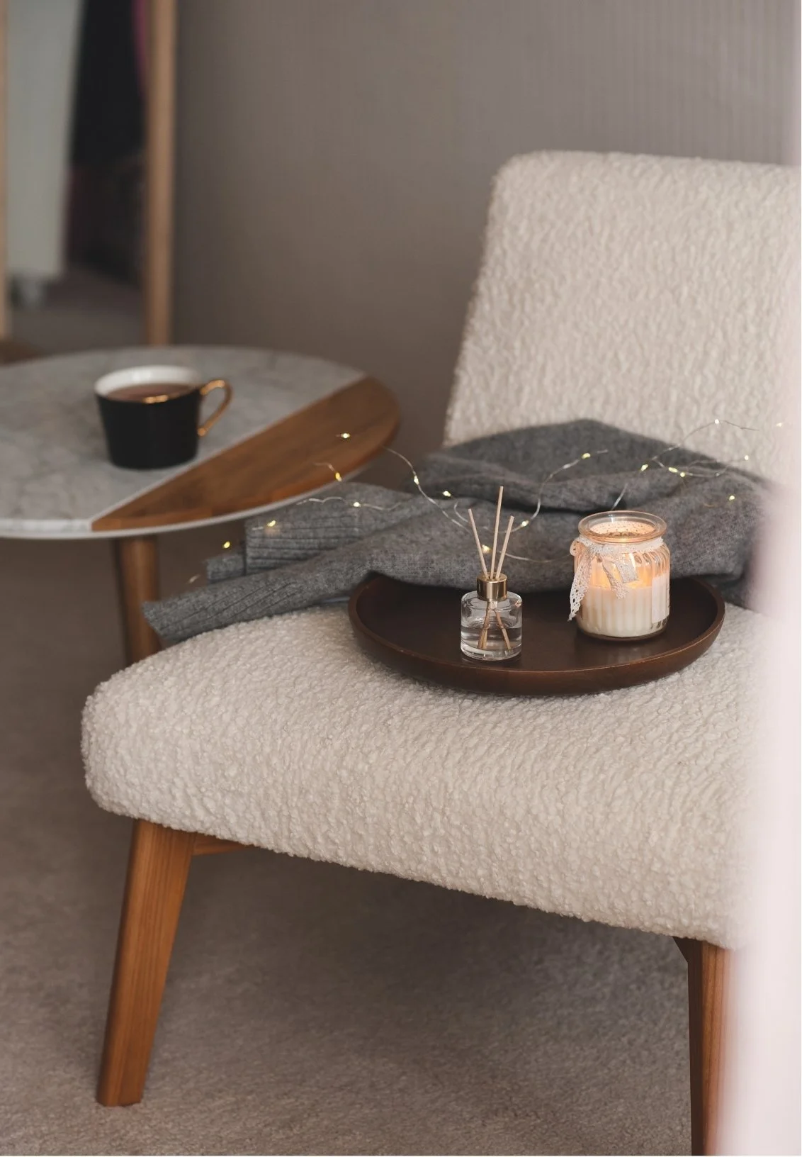 Cream-colored upholstered chair with wooden legs, gray blanket, and a brown tray holding a candle and reed diffuser, with a side table featuring a black cup and saucer in the background.