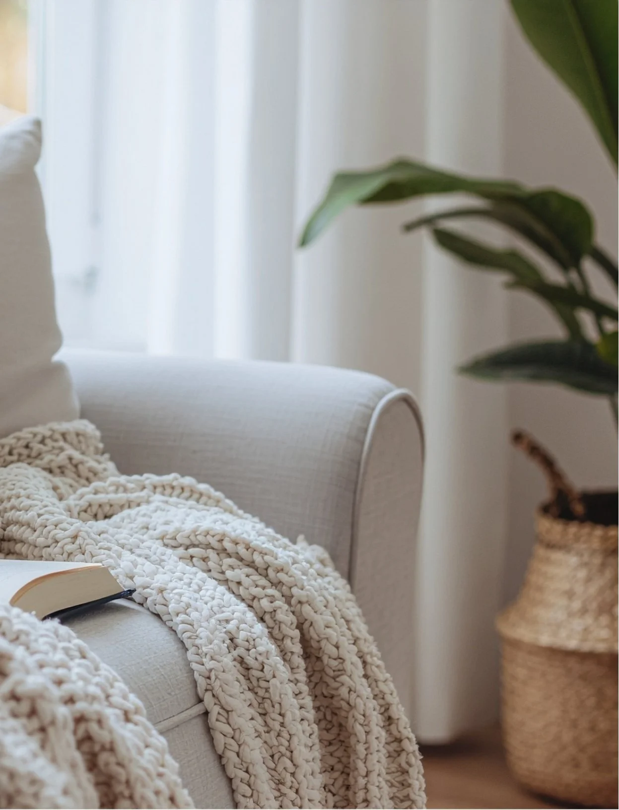 Cozy living room with a cream-colored couch, chunky knit blanket, open book, large plant, and woven basket in the background.