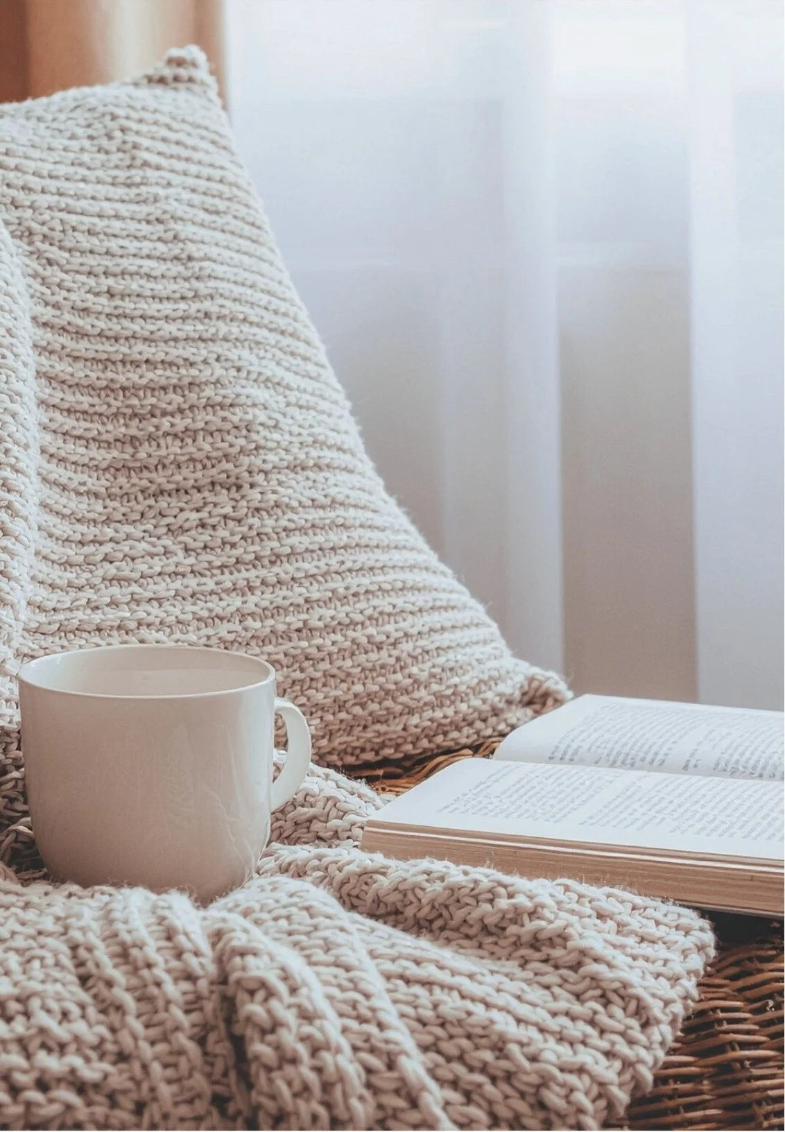 A cozy scene with an open book and a white coffee mug set on a knitted blanket, with a pillow in the background.