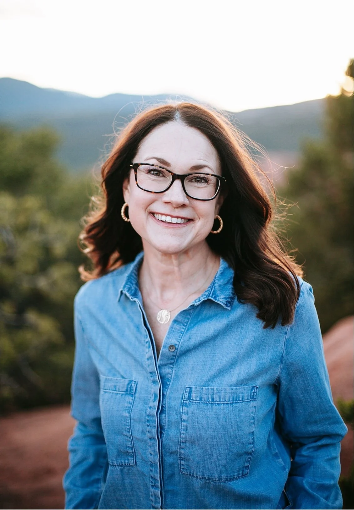 Colorado Springs therapist Celia Sugg smiles with mountains and greenery in the background during sunset.