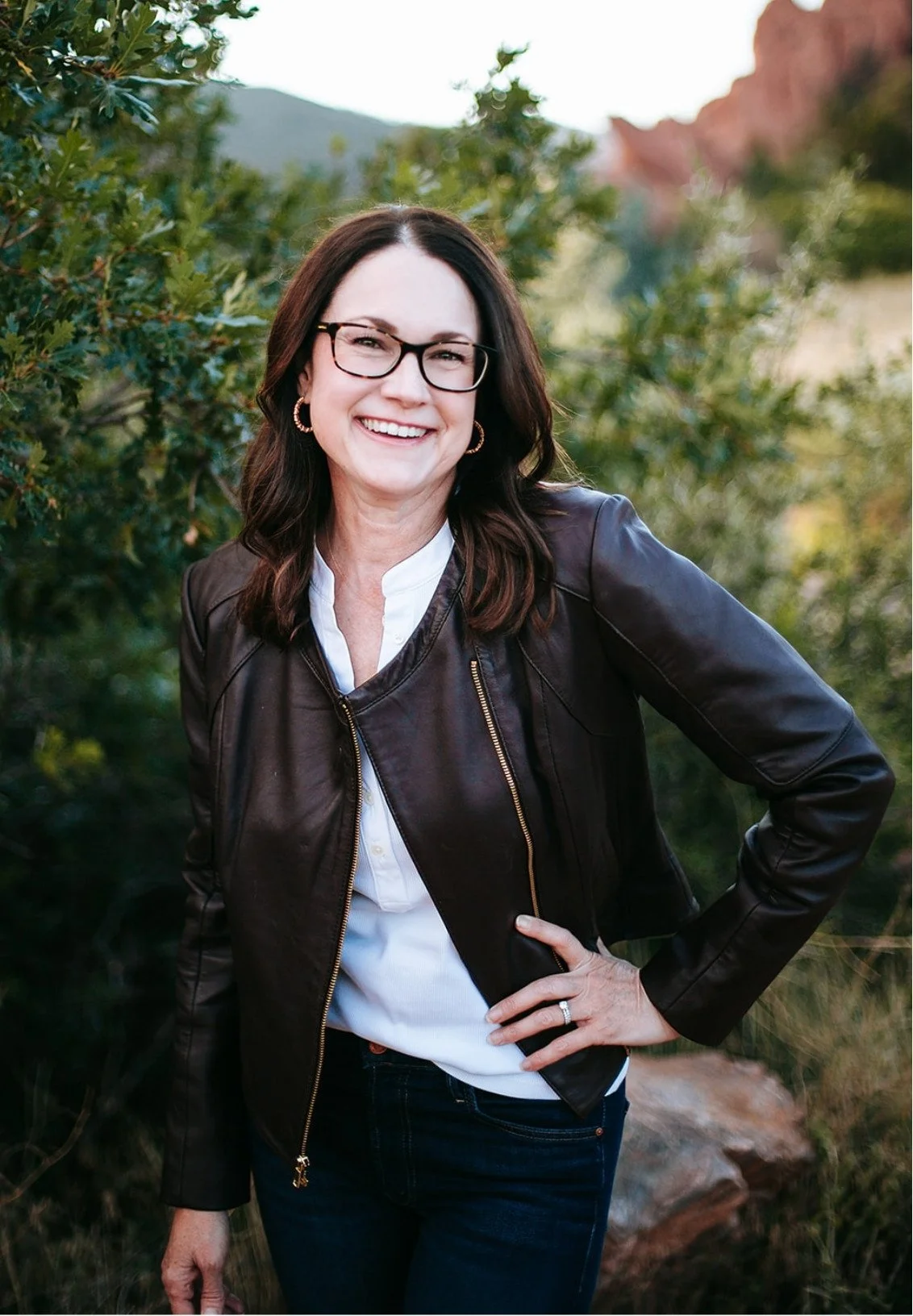 Celia Sugg, a therapist in Colorado Springs, smiles with a natural, green, and rocky background.