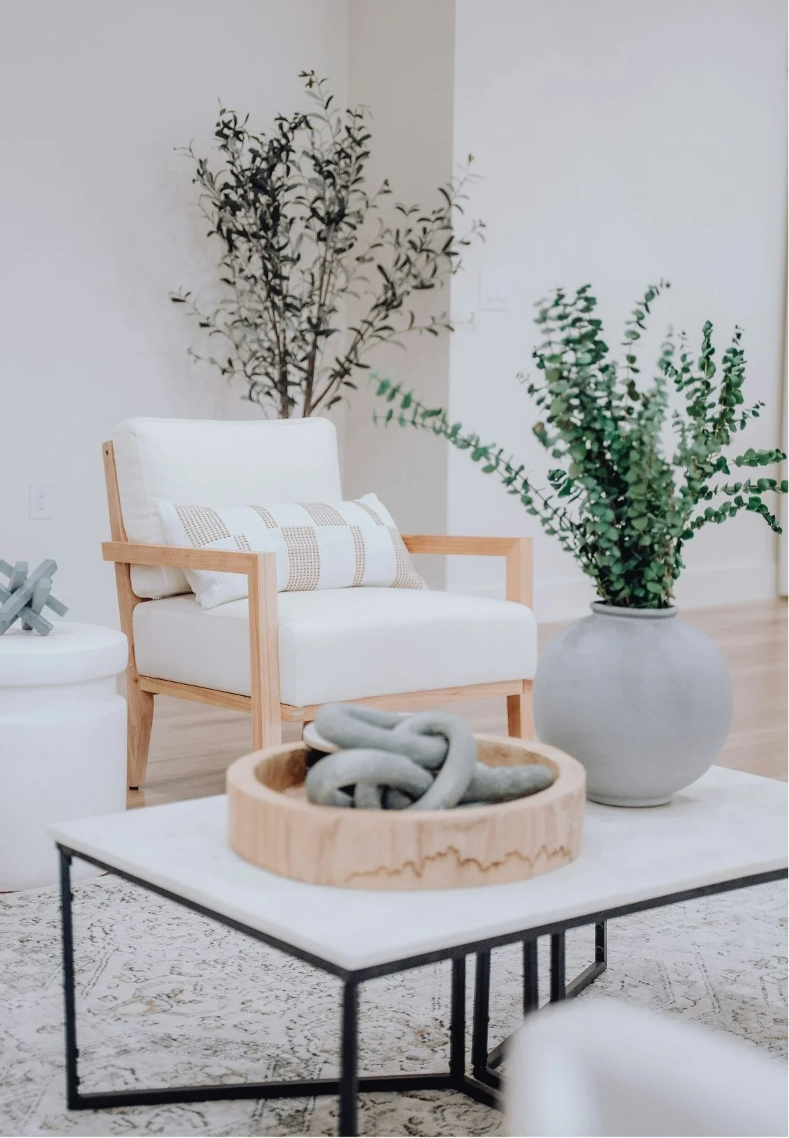 Minimalist living room with a white armchair with light wood arms and a decorative pillow, a large potted green plant in a gray vase, and a black metal coffee table with a wooden tray holding decorative stones.