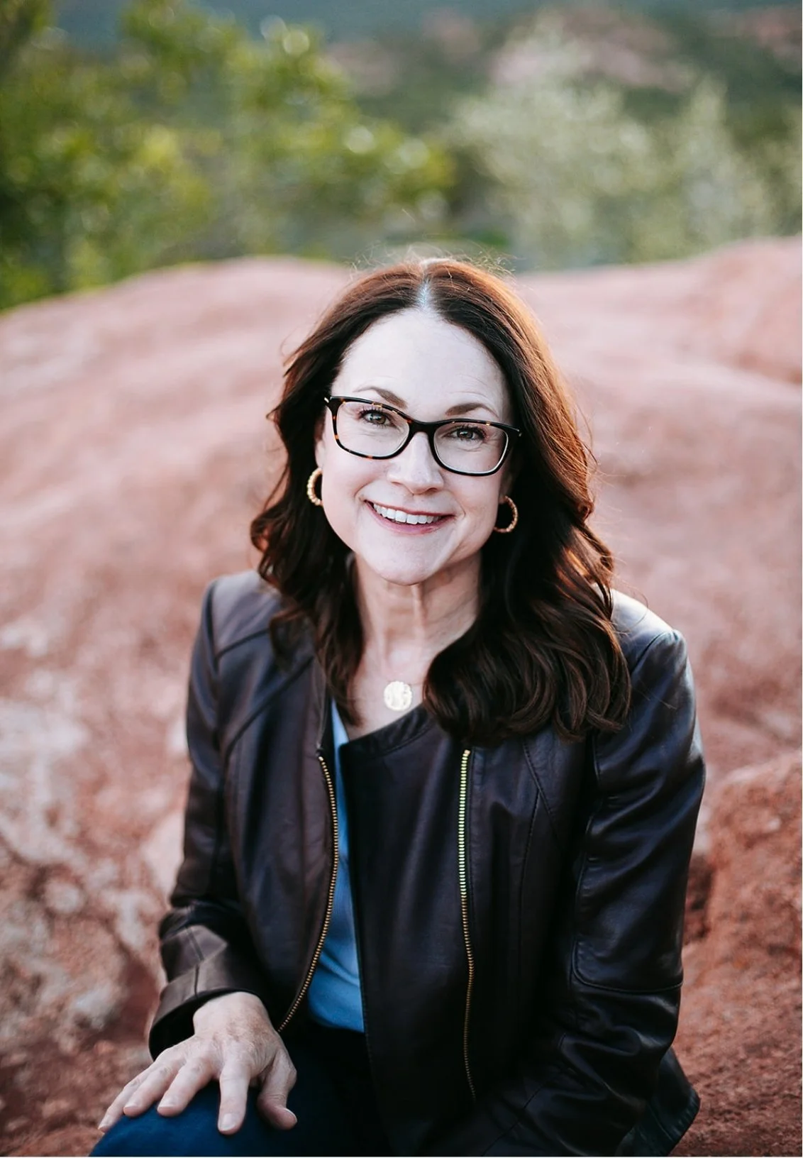 Celia Sugg, Colorado Springs anxiety therapist, sitting outdoors on a large rock with blurred greenery in the background, smiling at the camera.