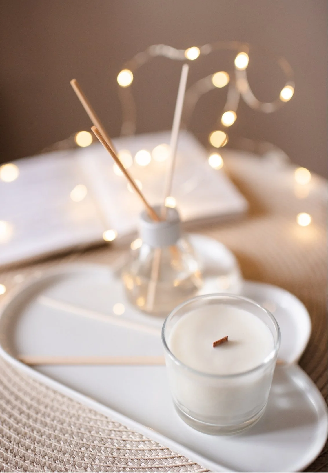 A white candle in a glass holder on a decorative tray with reed diffusers and string lights in the background.