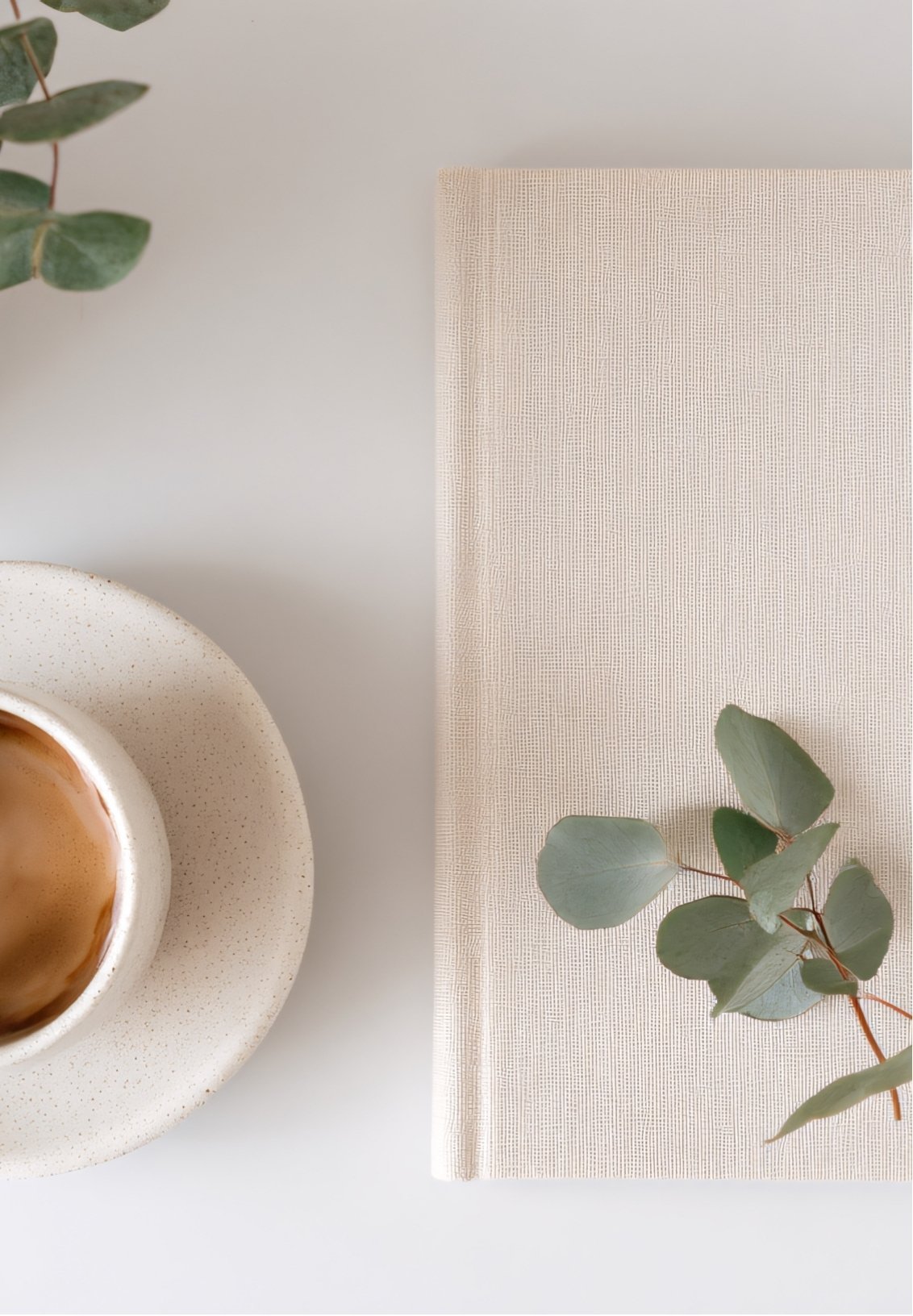 A white speckled cup of coffee on a matching saucer, a beige textured cloth, and a eucalyptus branch on a white surface.