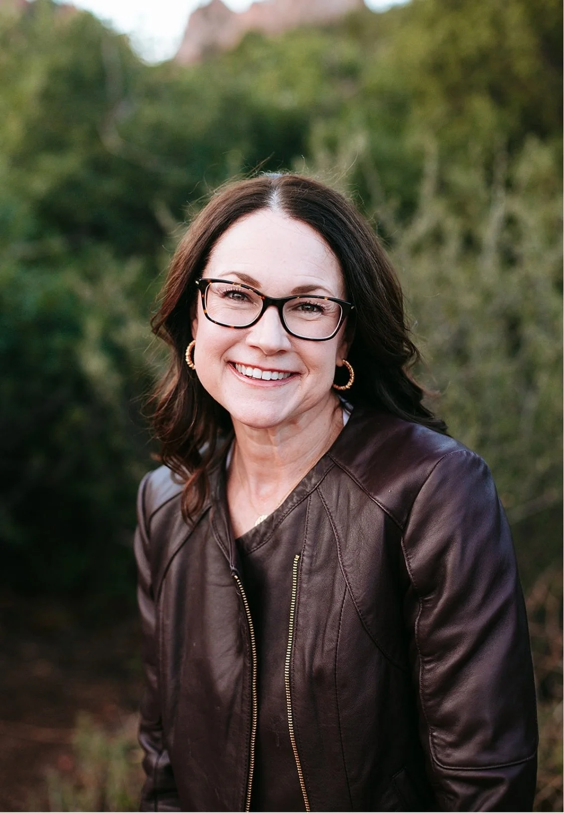 Celia Sugg, therapist in Colorado Springs, smiling at the camera, wearing glasses, hoop earrings, and a brown leather jacket, with a blurred natural green background.