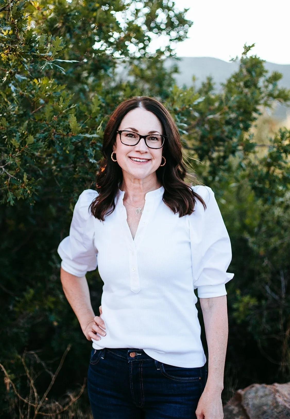 Colorado Springs therapist Celia Sugg smiling outdoors in front of green shrubbery, wearing a white blouse and dark jeans.