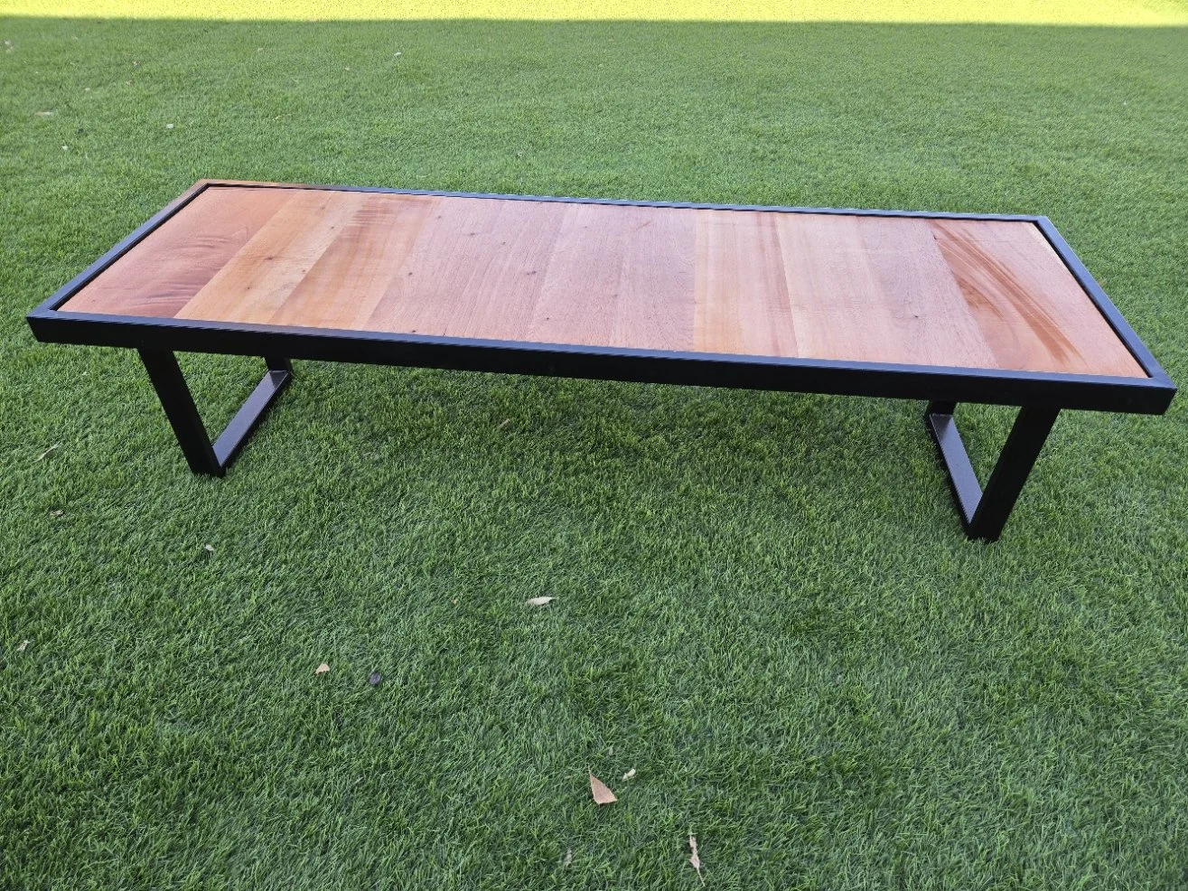 A rectangular coffee table with a cedar wooden top and black metal frame, placed on green grass outdoors.