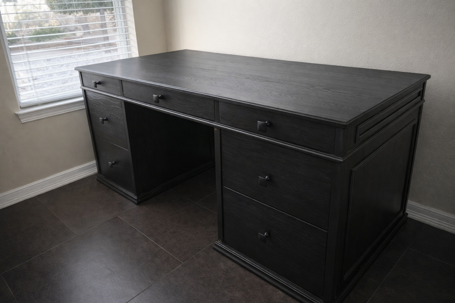 Black wooden desk with multiple drawers placed near a window with white blinds in a room with beige walls and dark tiled floors.