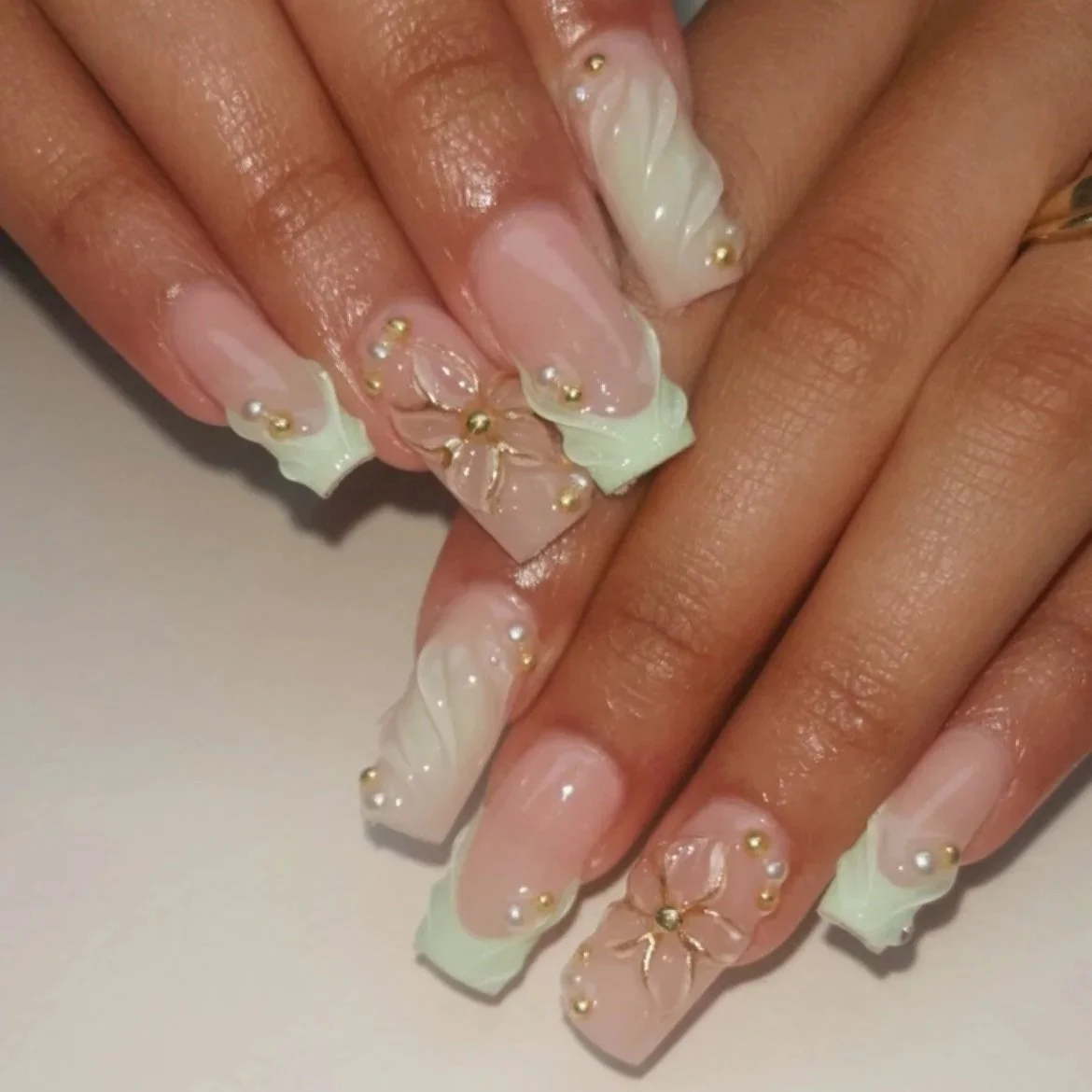 Close-up of a hand with elaborate long acrylic nails featuring 3D flower designs, pearls, and gold accents, in pastel pink, white, and beige colors.