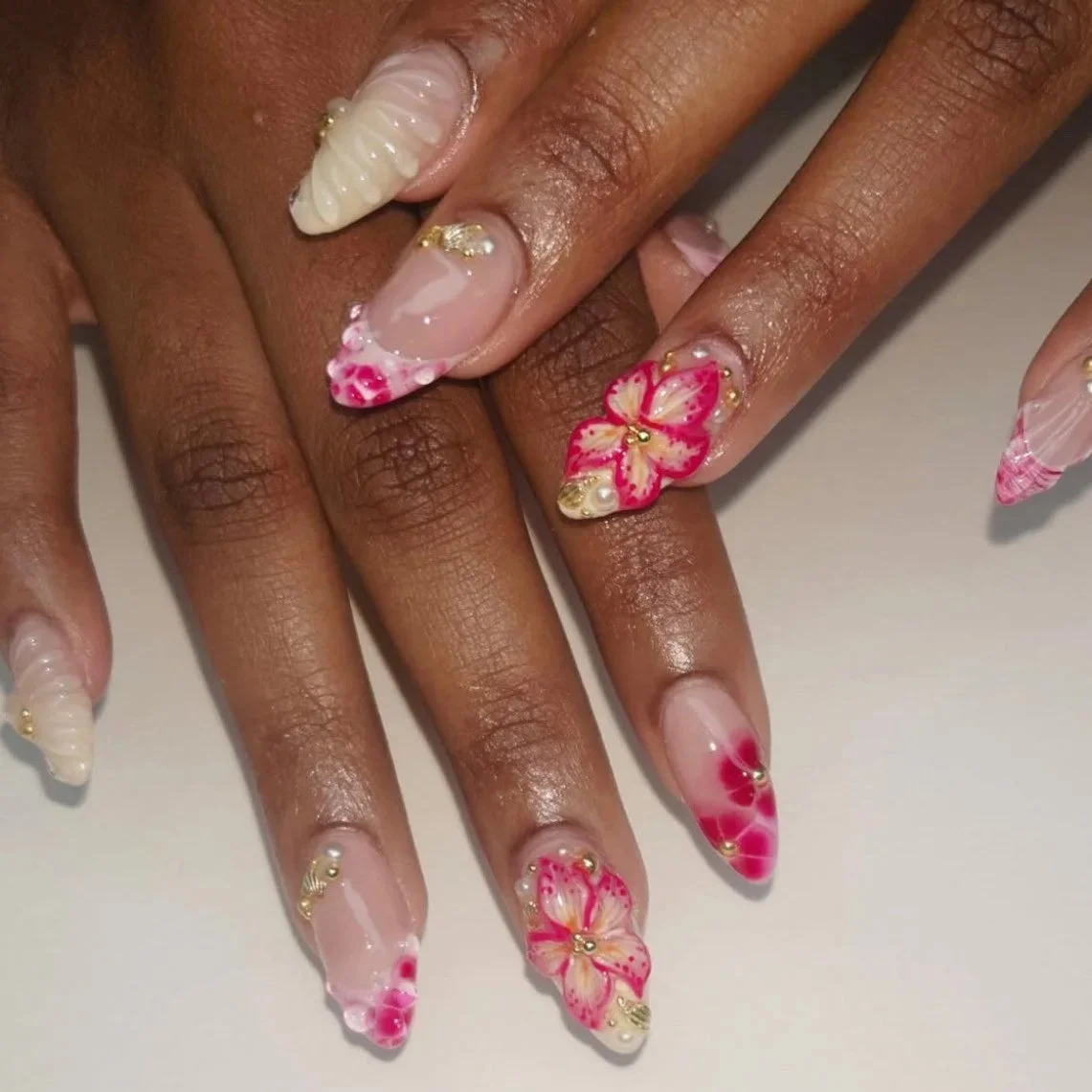 Close-up of a person's hand with elaborately decorated nail art featuring pink and cream flower designs, rhinestones, and gold accents.
