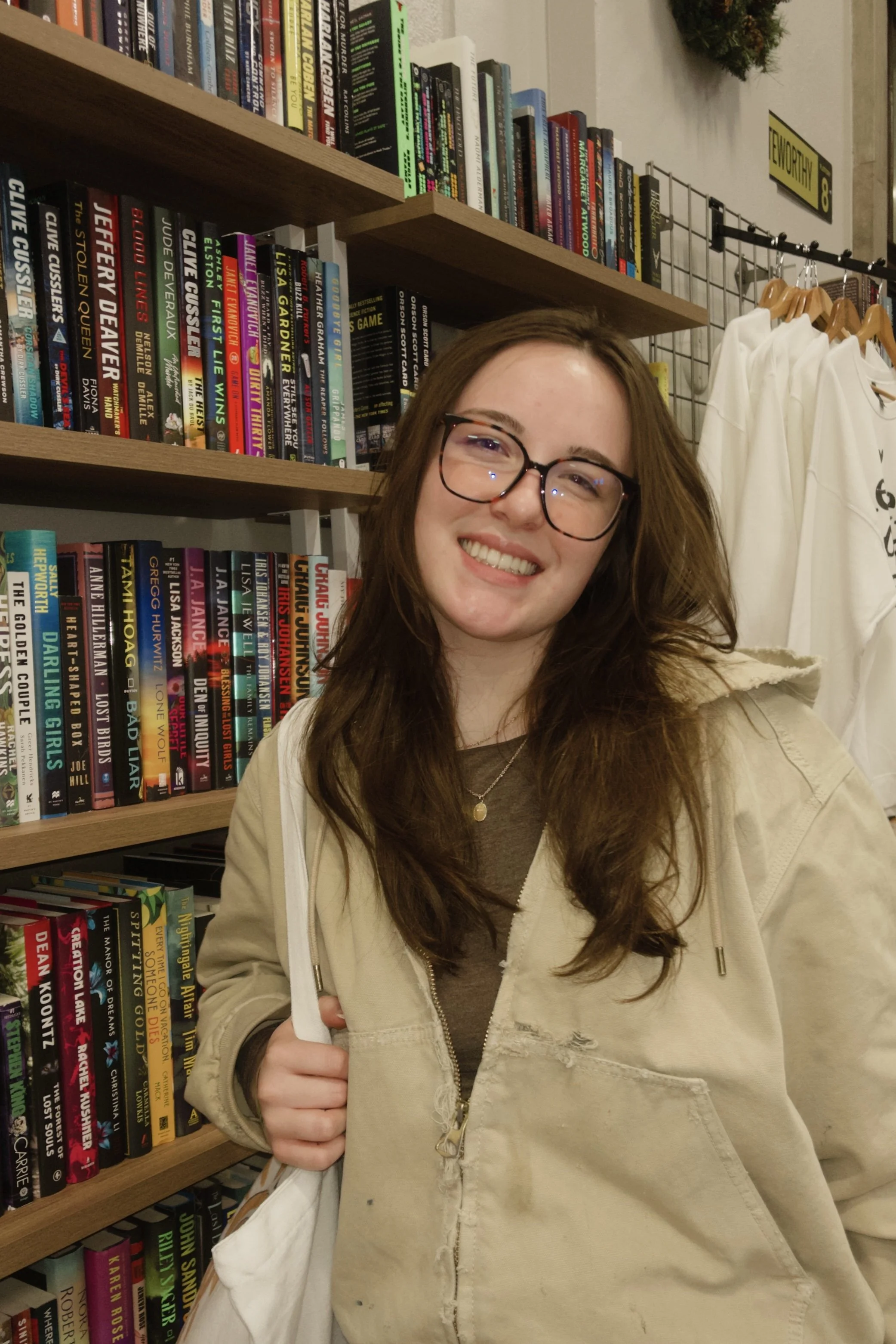 A young woman with glasses and long brown hair smiling, standing in front of a bookshelf filled with colorful books, wearing a beige jacket and a necklace.