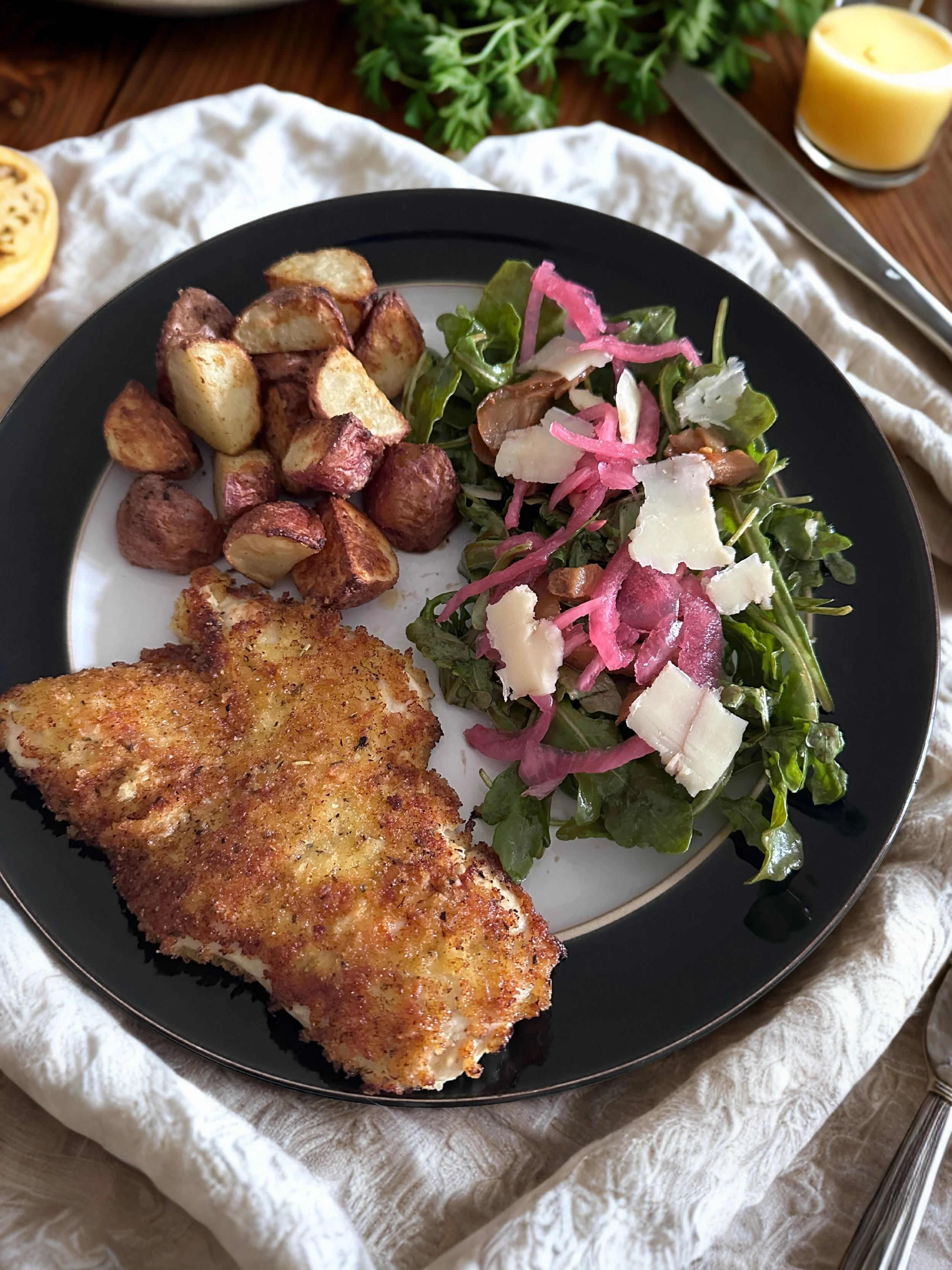 Plate with fried fish fillet, roasted potatoes, and a mixed green salad with cheese, pickled onions, and dressing, on a black and white plate.