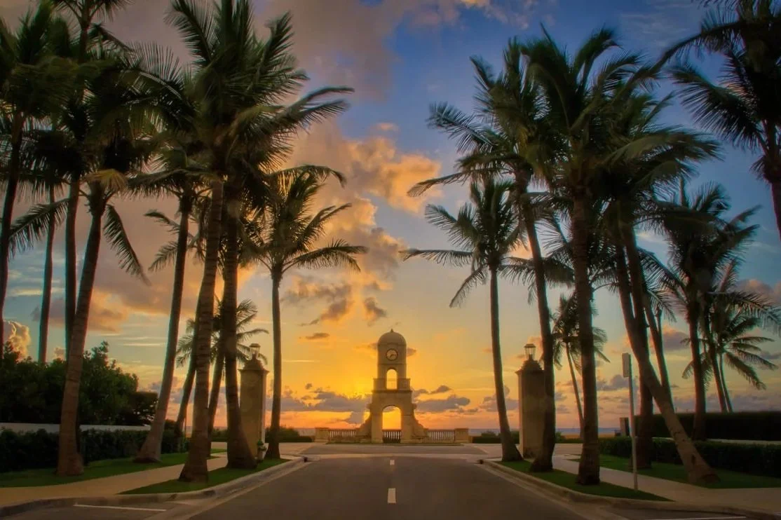 Iconic Worth Ave clock tower at sunset framed by palm trees along a scenic road in Palm Beach Florida