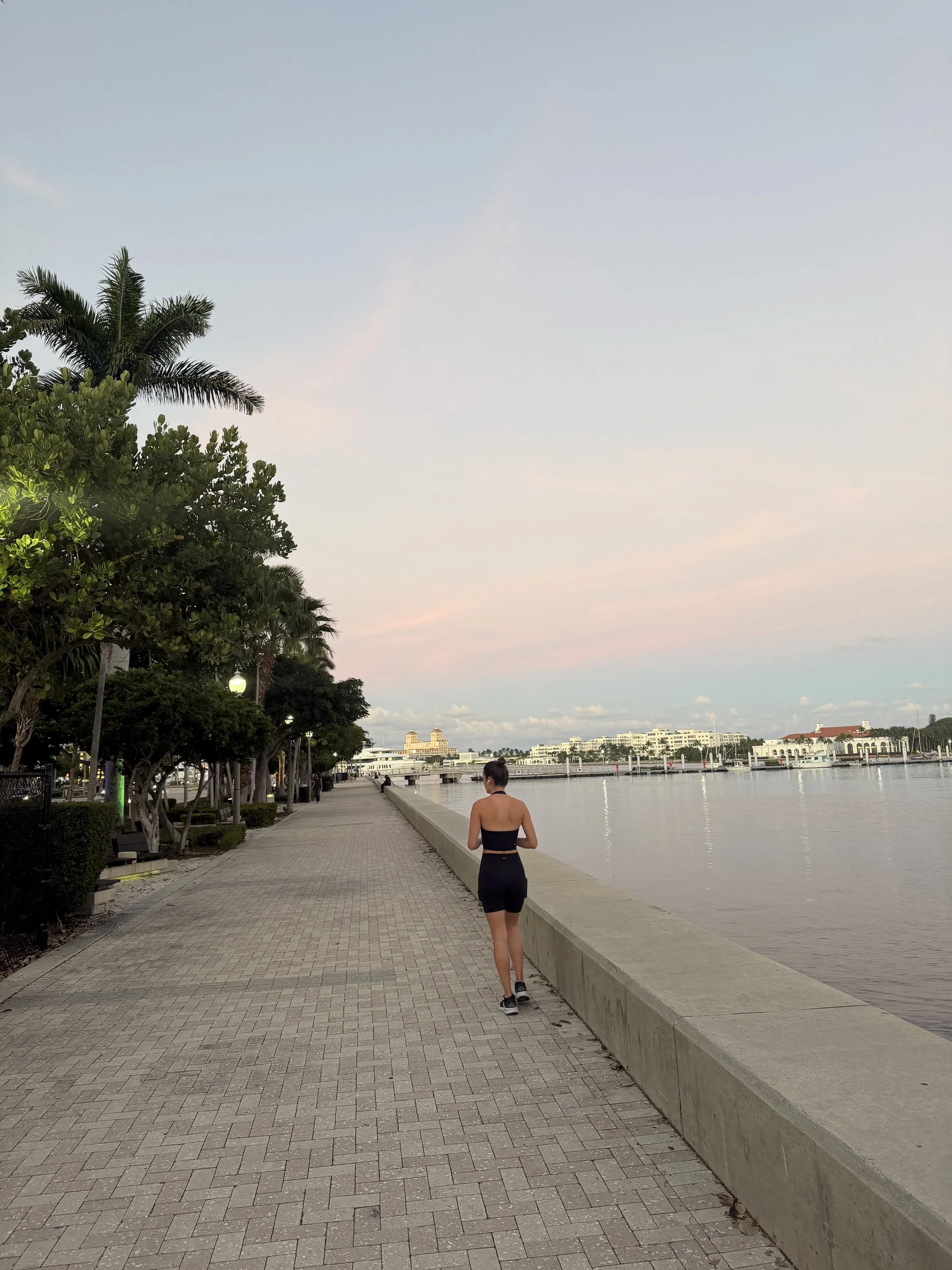 A woman jogging along a waterfront paved walkway at sunrise or sunset, with trees on the left and water on the right, across from buildings and boats in the distance.