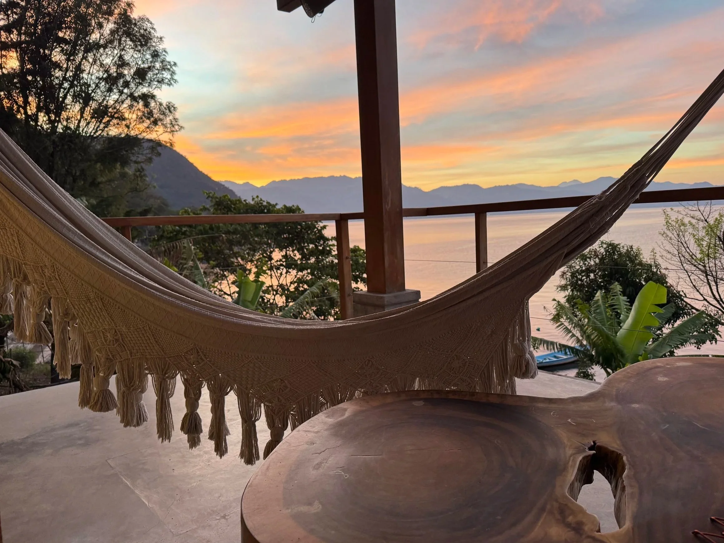 Hammock hanging on a porch overlooking a lake and mountain range at sunset.