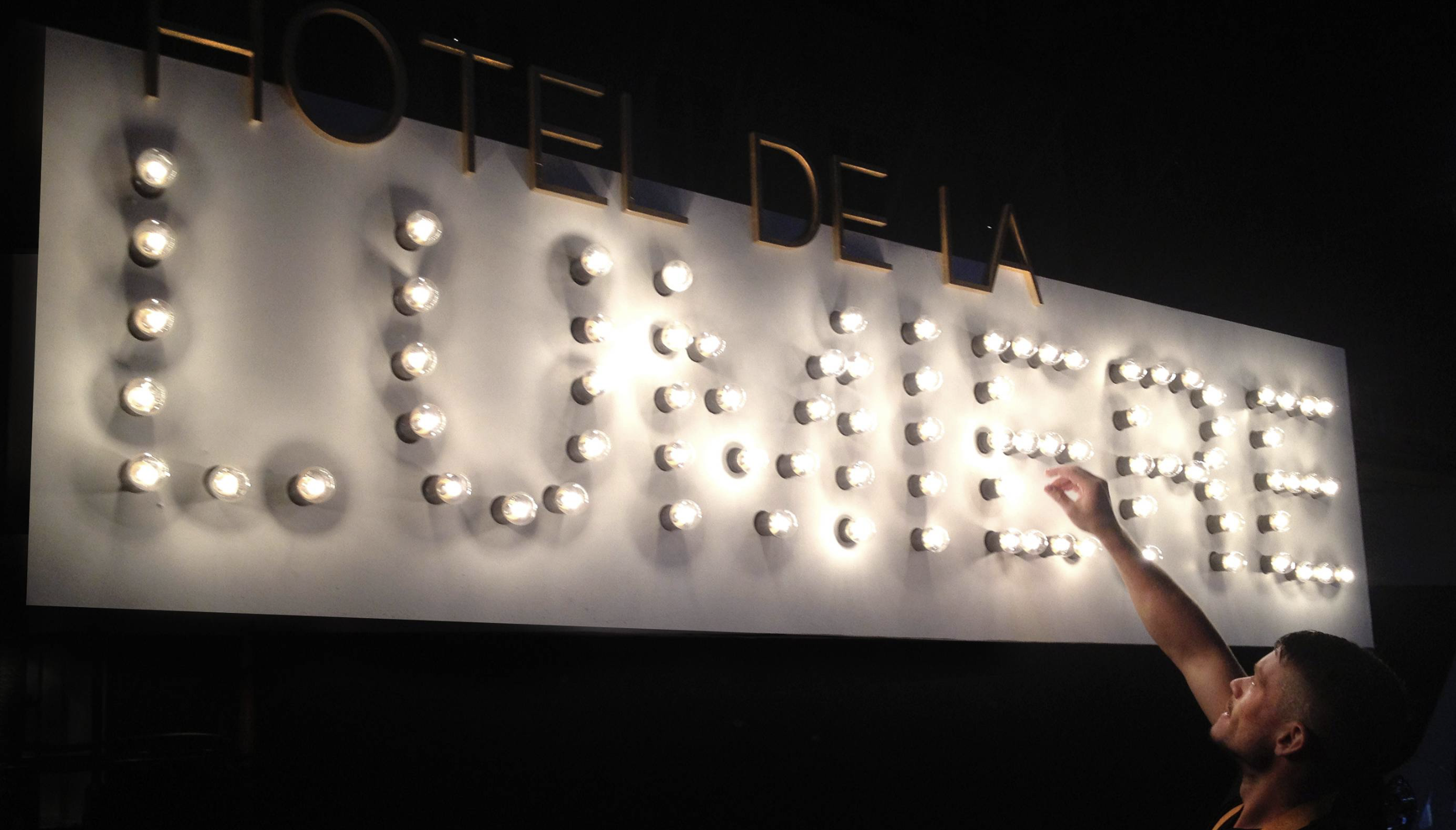 A person reaching towards a large illuminated sign with the word 'LOVE' made of light bulbs, mounted on a white background, with the word 'HOTELDELA' partially visible at the top.