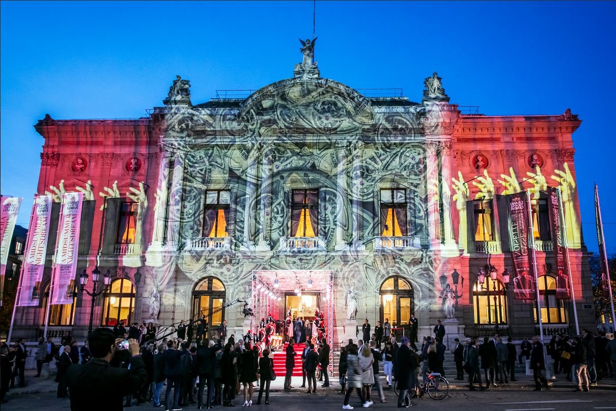 A historic building is illuminated with colorful lights and projection displays during an event at night. People are gathered outside, some taking photos, while a few are entering through the main entrance.