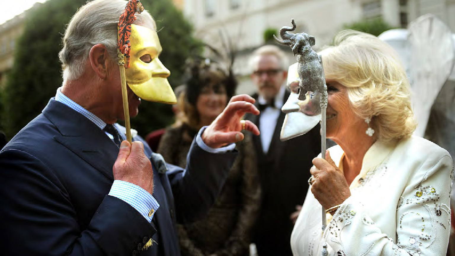 Two people wearing masks, one resembling a yellow face and the other resembling a white face with a small animal on top, are interacting at an outdoor event with onlookers in the background.