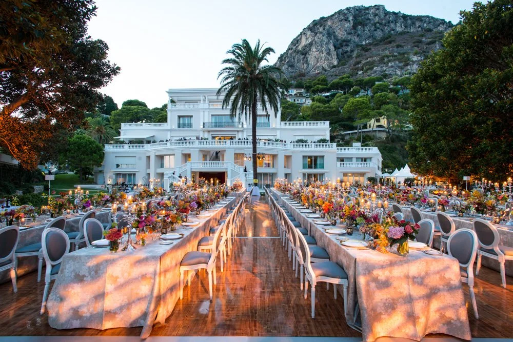 Elegant outdoor wedding reception setup with long banquet tables adorned with colorful floral centerpieces and candlelit decorations, set against a large white mansion and lush green hills with mountains in the background.