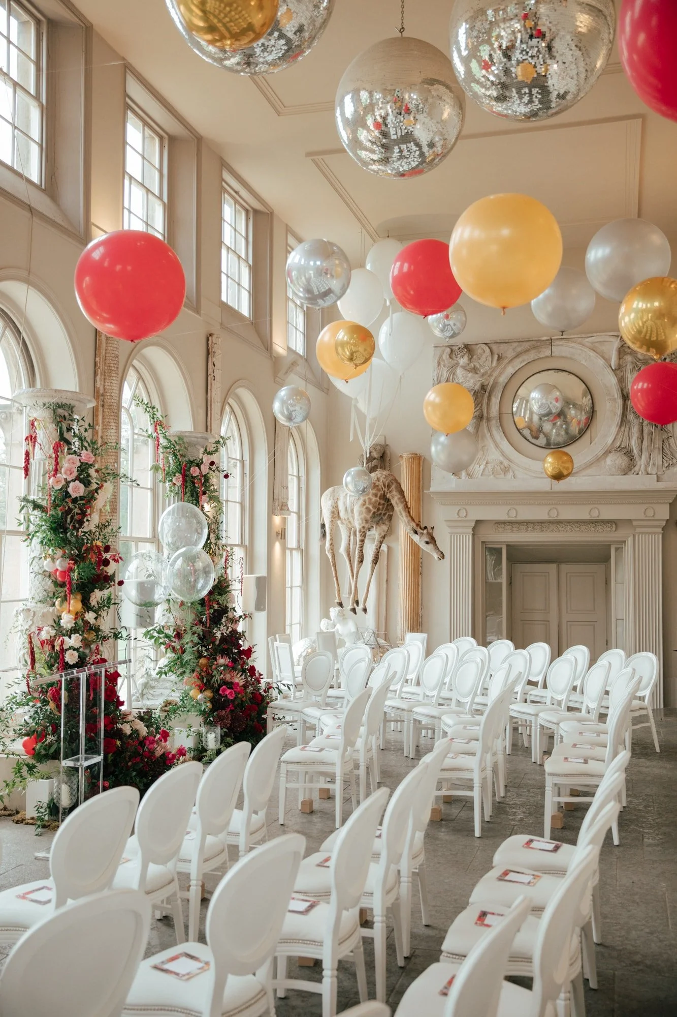 Decorated room with white chairs, floral arrangements, balloons, and a giraffe statue, likely set up for a celebration or event.