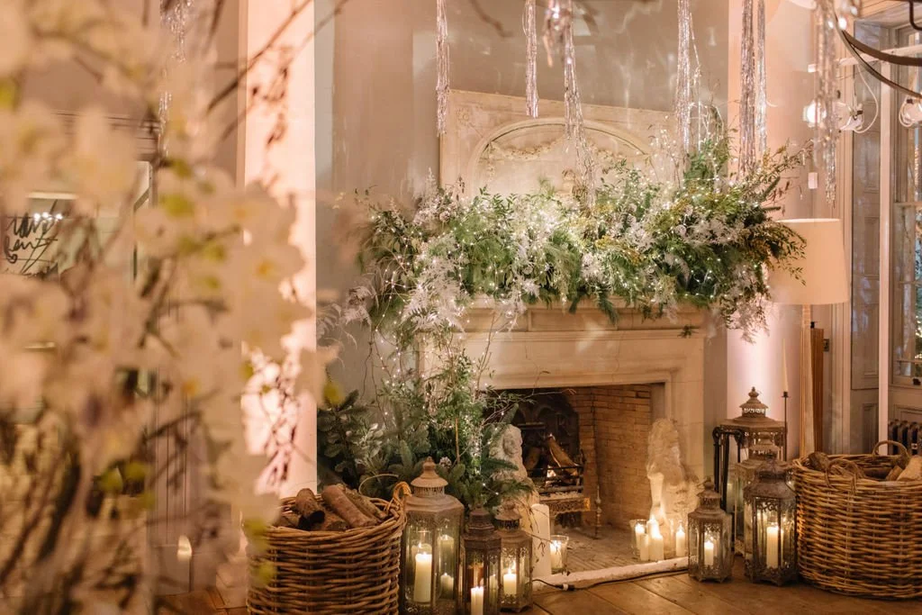 Decorated fireplace mantle with floral arrangements, string lights, and candles in lanterns, creating a cozy and elegant setting.