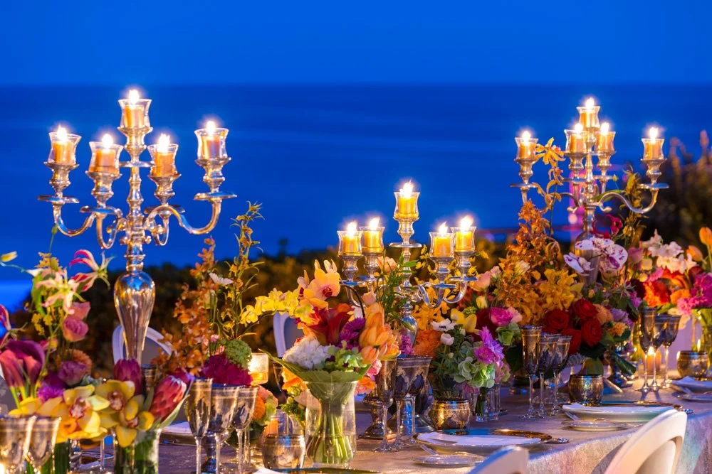 Elegant outdoor dinner table with multiple gold candelabras with lit candles, colorful floral arrangements, and glassware, set against a backdrop of the ocean during twilight.