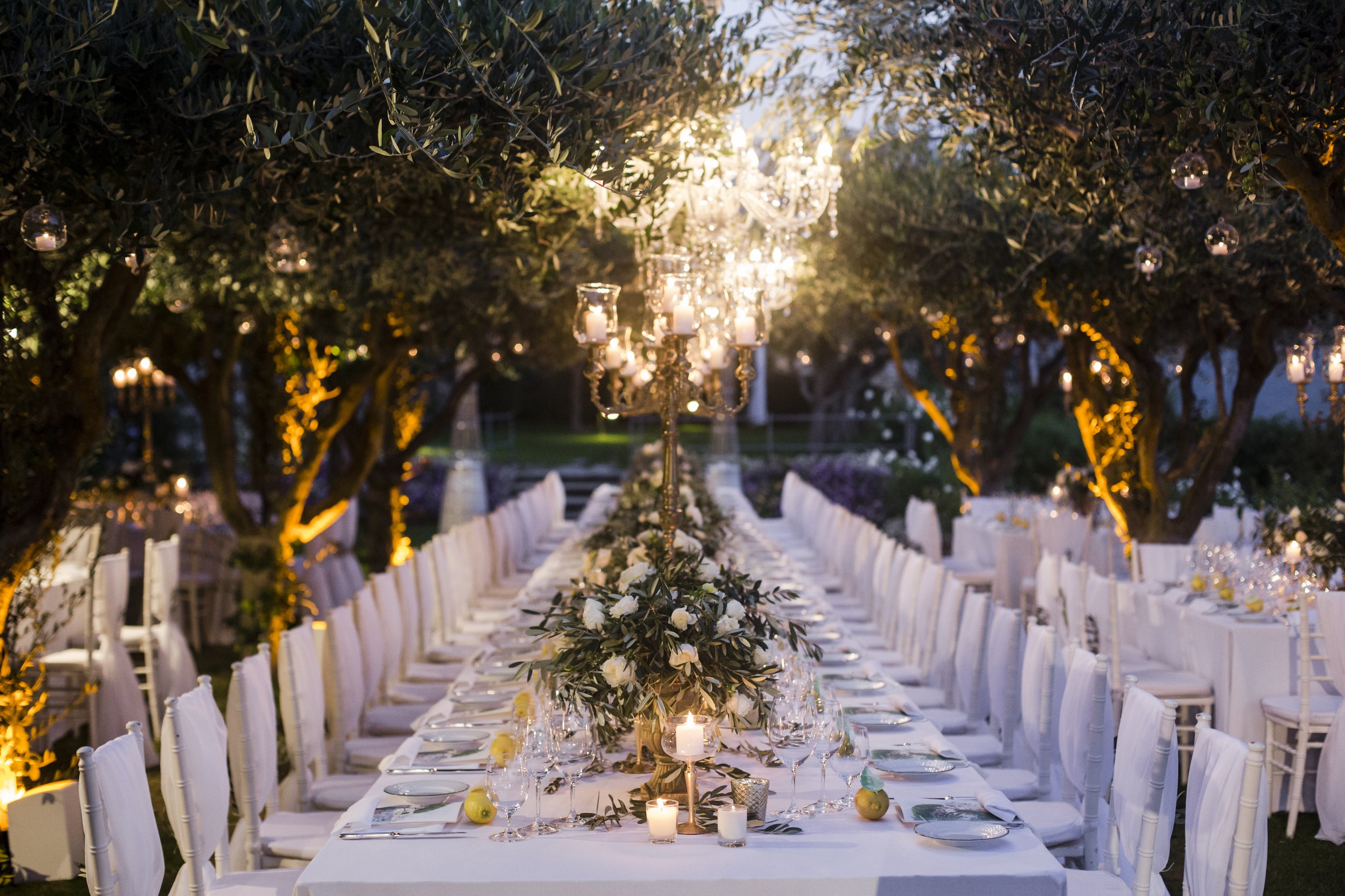 A long outdoor dining table decorated with flowers, candles, and elegant tableware is set under trees with hanging lights and lanterns, creating a festive evening atmosphere.