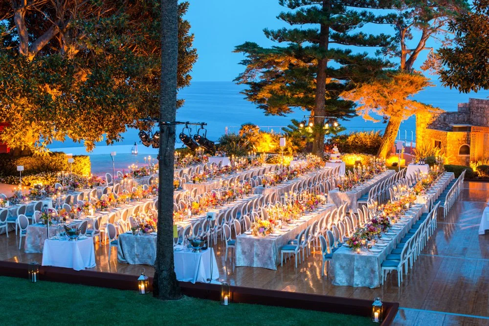An outdoor dinner event set with long tables arranged in a U-shape, decorated with floral centerpieces, near the coast with the ocean visible in the background, illuminated by warm lighting and lanterns.