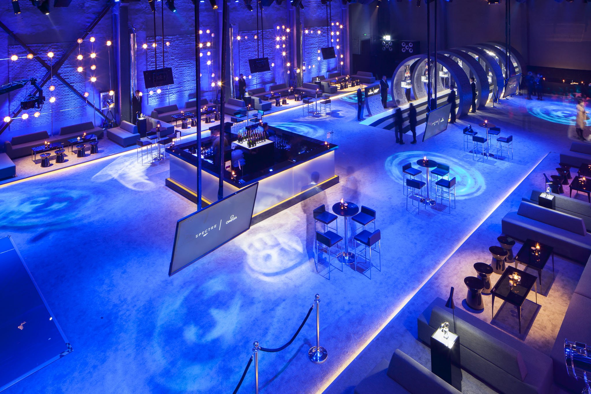 Aerial view of an upscale event space or lounge decorated with blue lighting, modern furniture, and illuminated circular motifs on the floor, with a bar at the center and several seating and standing areas.