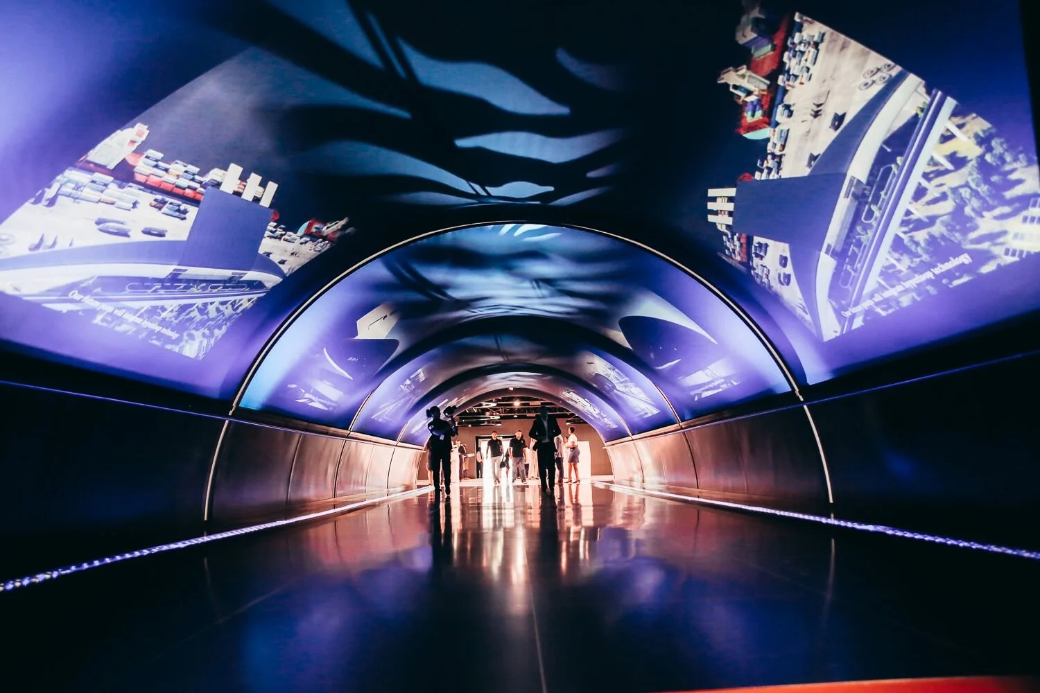 People walking through a futuristic tunnel with digital screens on the ceiling displaying cityscape images.