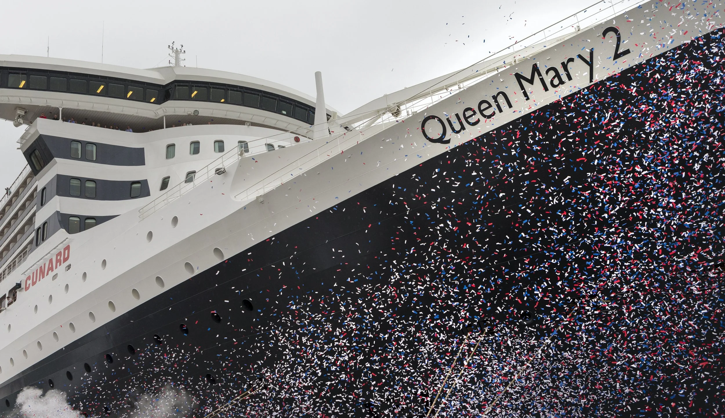 Close-up of the Queen Mary 2 ship's bow, decorated with red, white, and blue confetti, with the name 'Queen Mary 2' visible, during a celebration event.