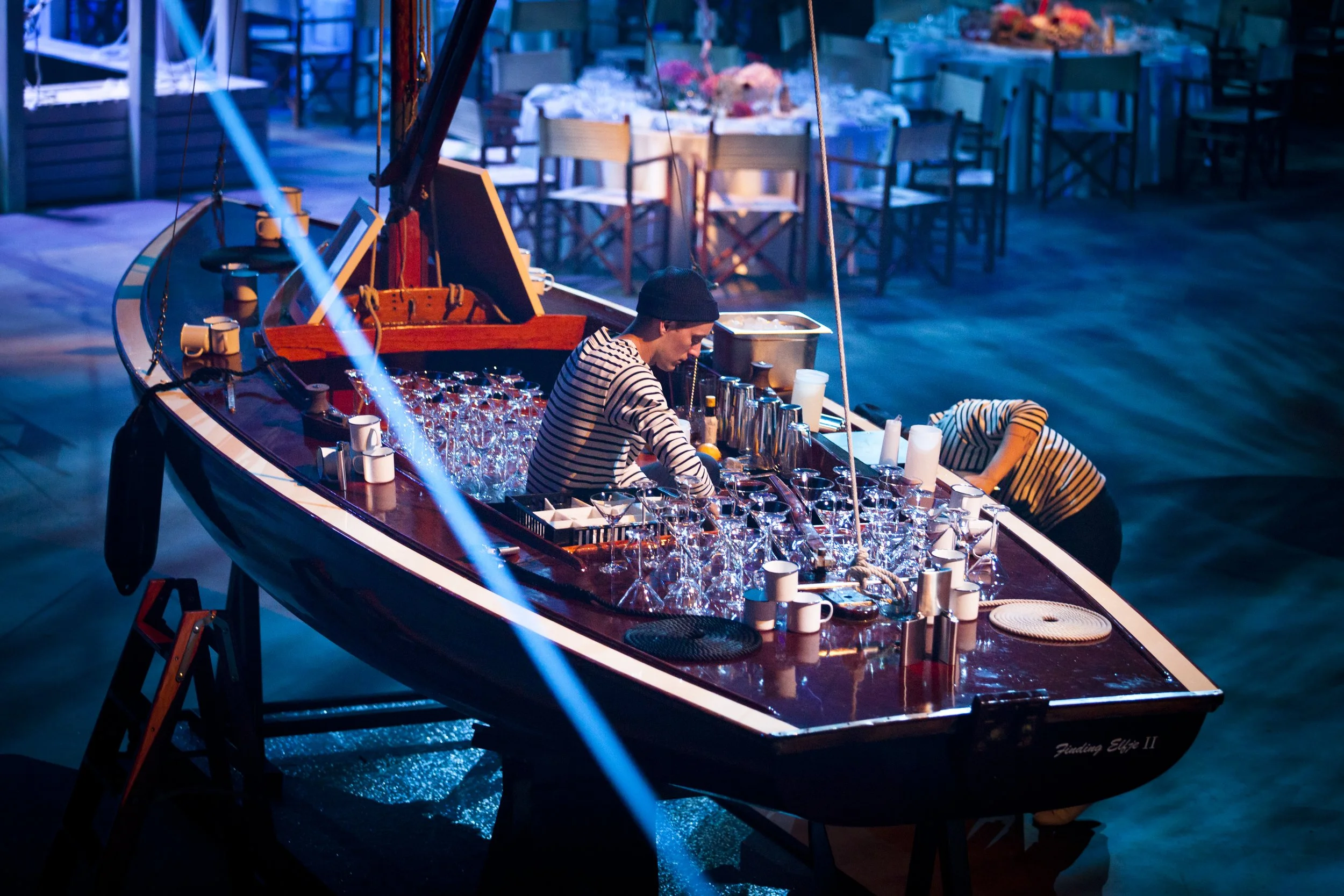 A person on a boat-shaped bar, wearing a black beanie and striped shirt, preparing drinks. The boat is inside a large indoor space with tables and chairs in the background, decorated with flowers.