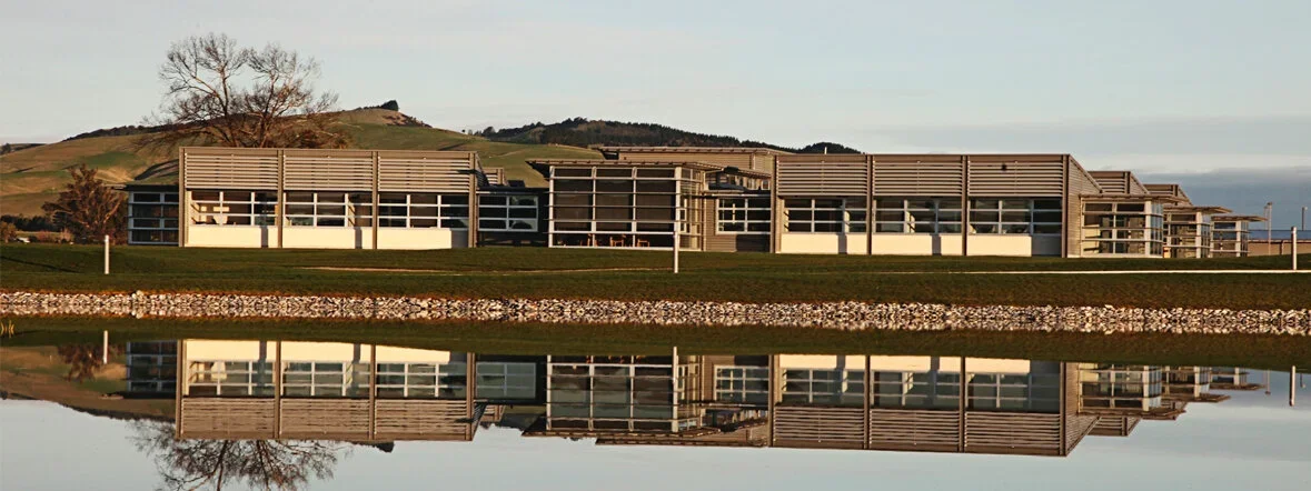 Modern building with horizontal wooden slats, large windows, grassy landscape, hills in background, and reflection in water.