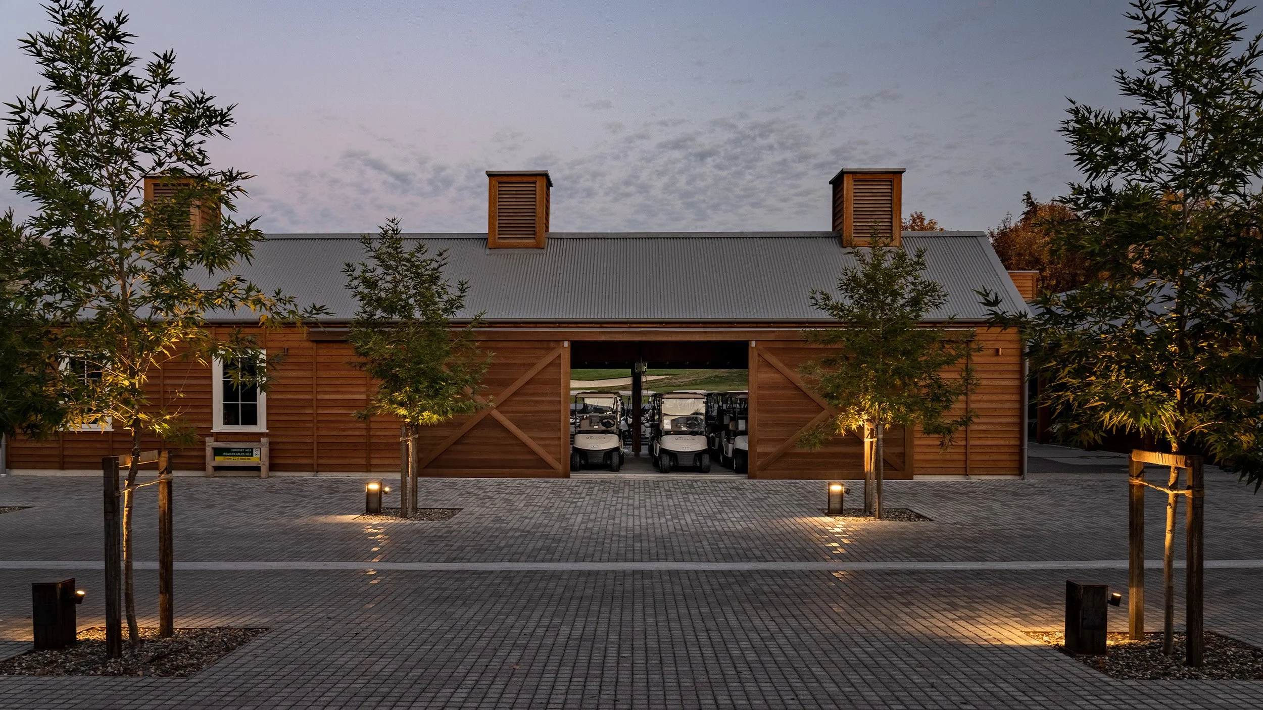 A wooden building with a metal roof, featuring two storybook-style chimneys. In front, there are small trees and cobblestone pathway, with golf carts parked inside the open doorway.
