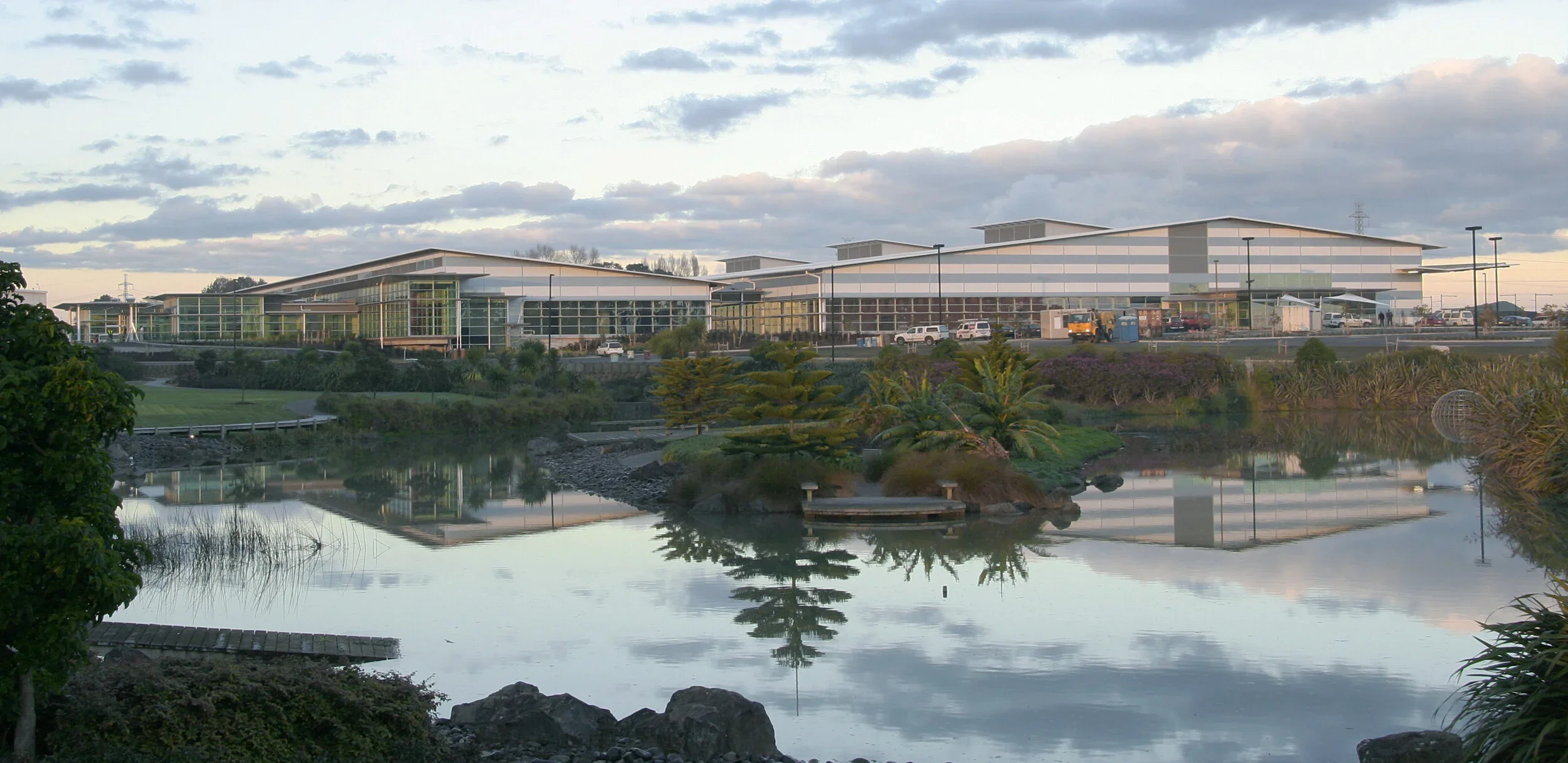 Modern building with glass and metal exterior situated near a pond with trees and bushes, reflecting the structure and sky.