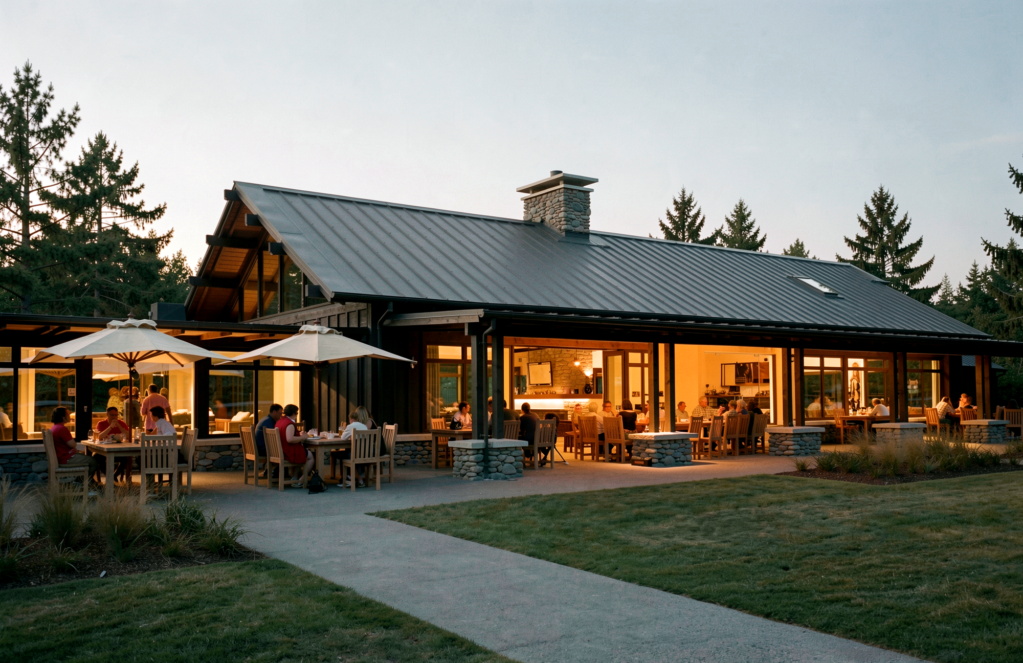 An outdoor restaurant or cafe with patrons dining at tables under umbrellas, adjacent to a modern house with large glass windows and a metal roof, set in a wooded area during evening.