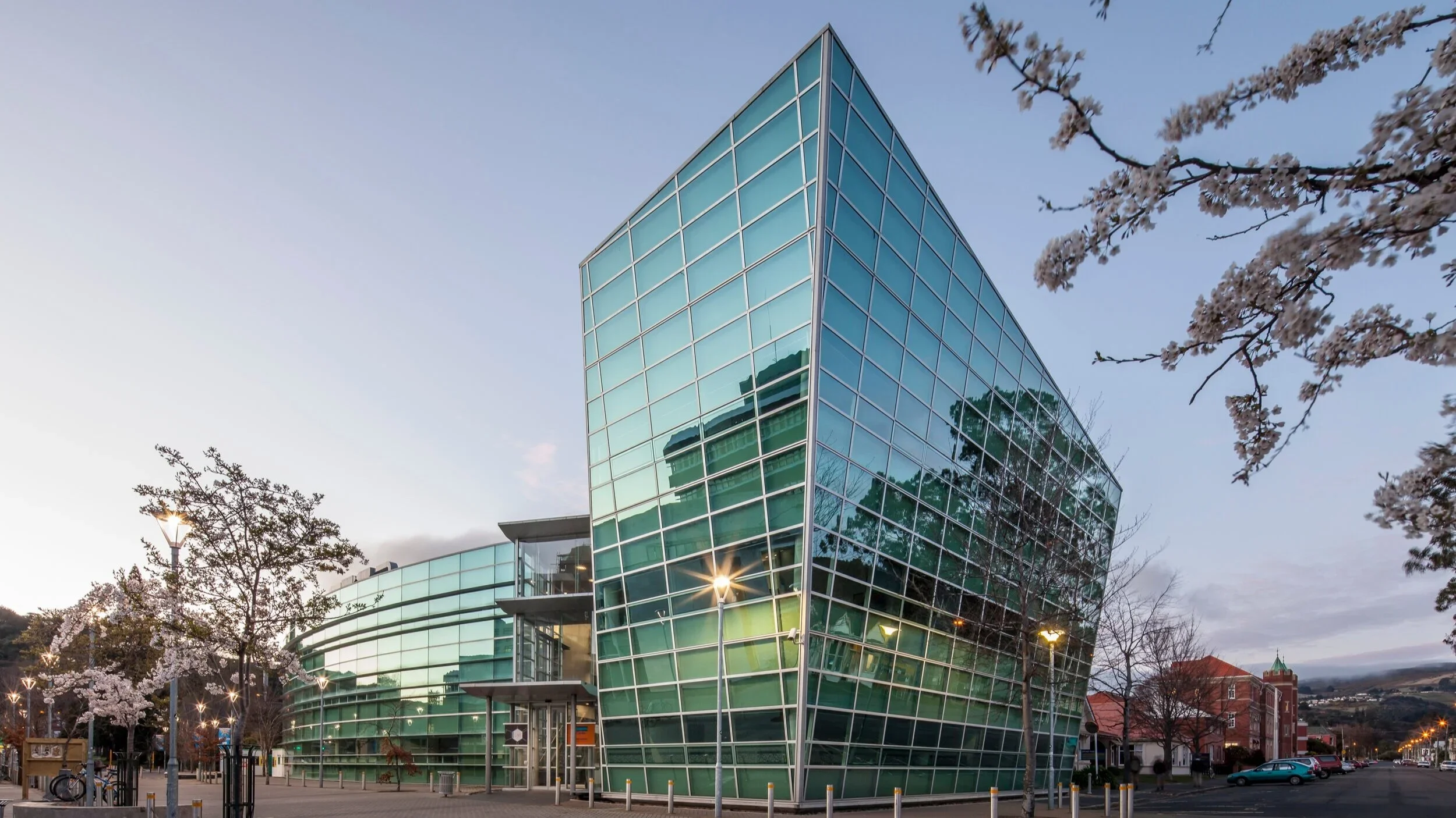 Modern glass building with a curved and angular design, surrounded by trees and streetlights, during dusk.