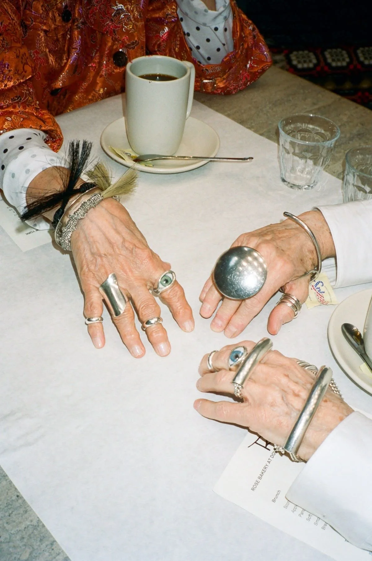 Three elderly people with jewelry and rings holding hands over a table with cups, water glasses, and silverware at a brunch setting.