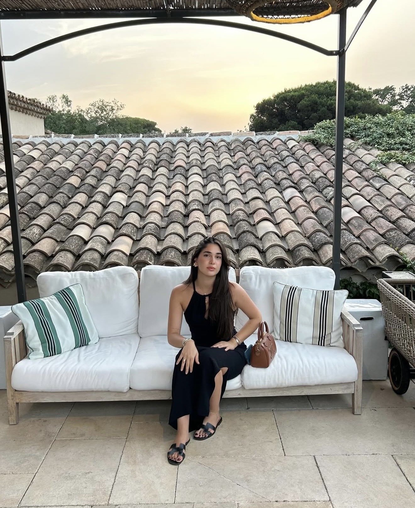 A woman in a black dress sitting on a white outdoor sofa with striped pillows, in front of a tiled roof, during sunset or early evening.